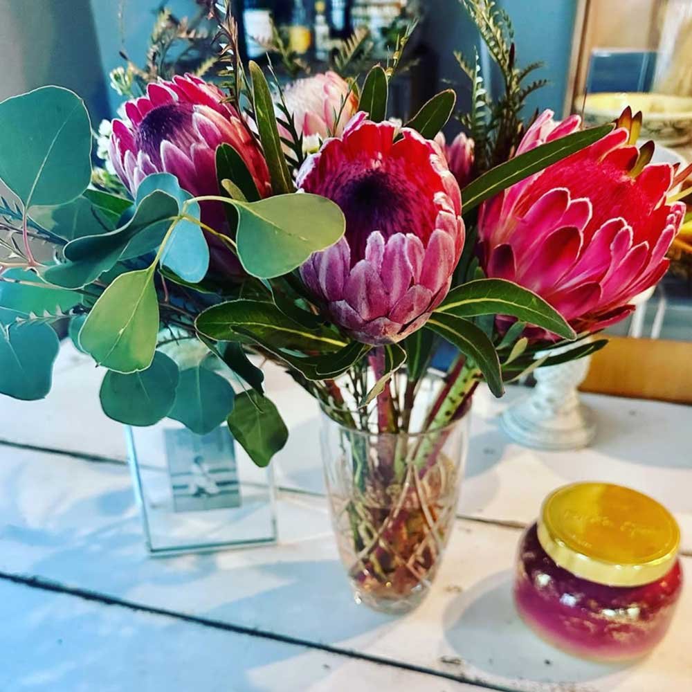 A bouquet of pink and purple protea flowers with green foliage in a clear glass vase on a white table in hair studio, with a small yellow-capped jar nearby.