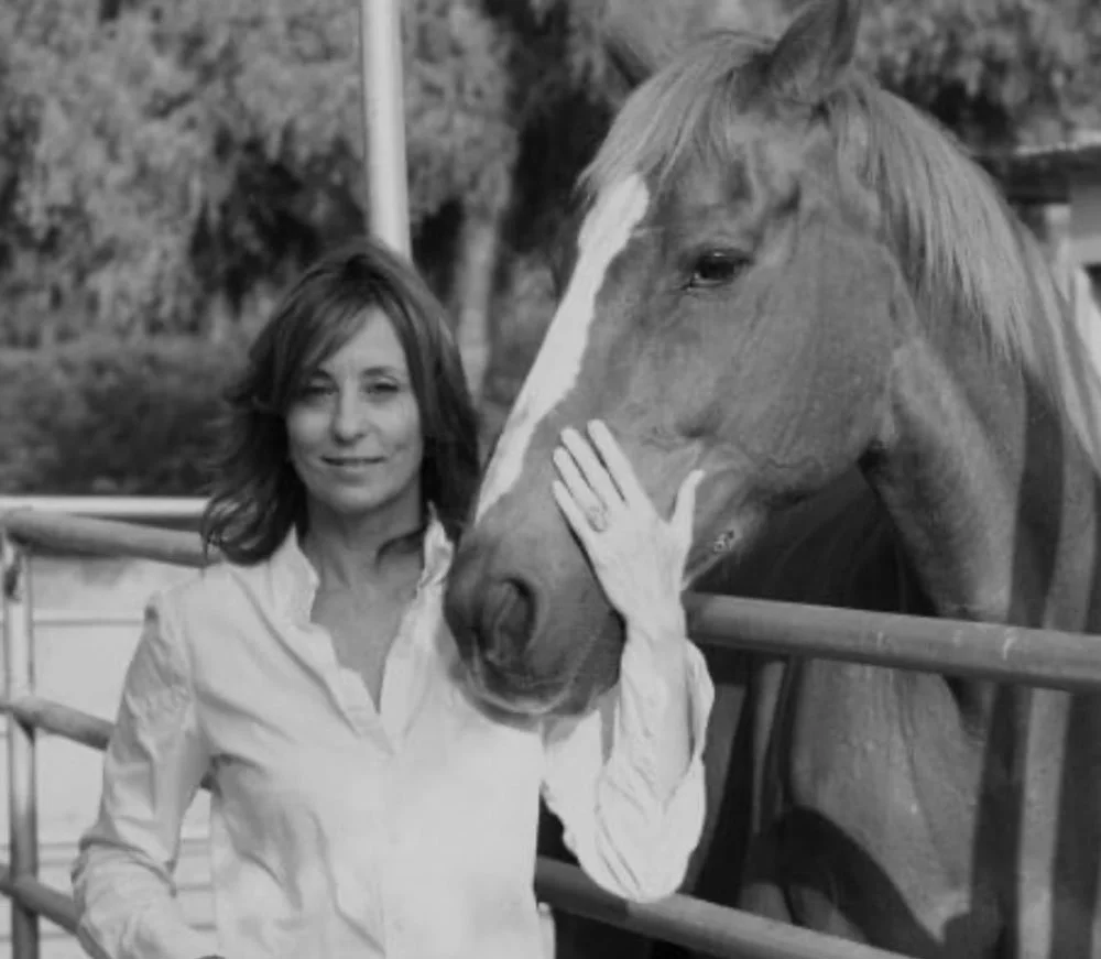 A woman with shoulder-length hair and a white shirt standing next to a horse with her hand touching its face, behind a fence. In the background, trees and outdoor setting.