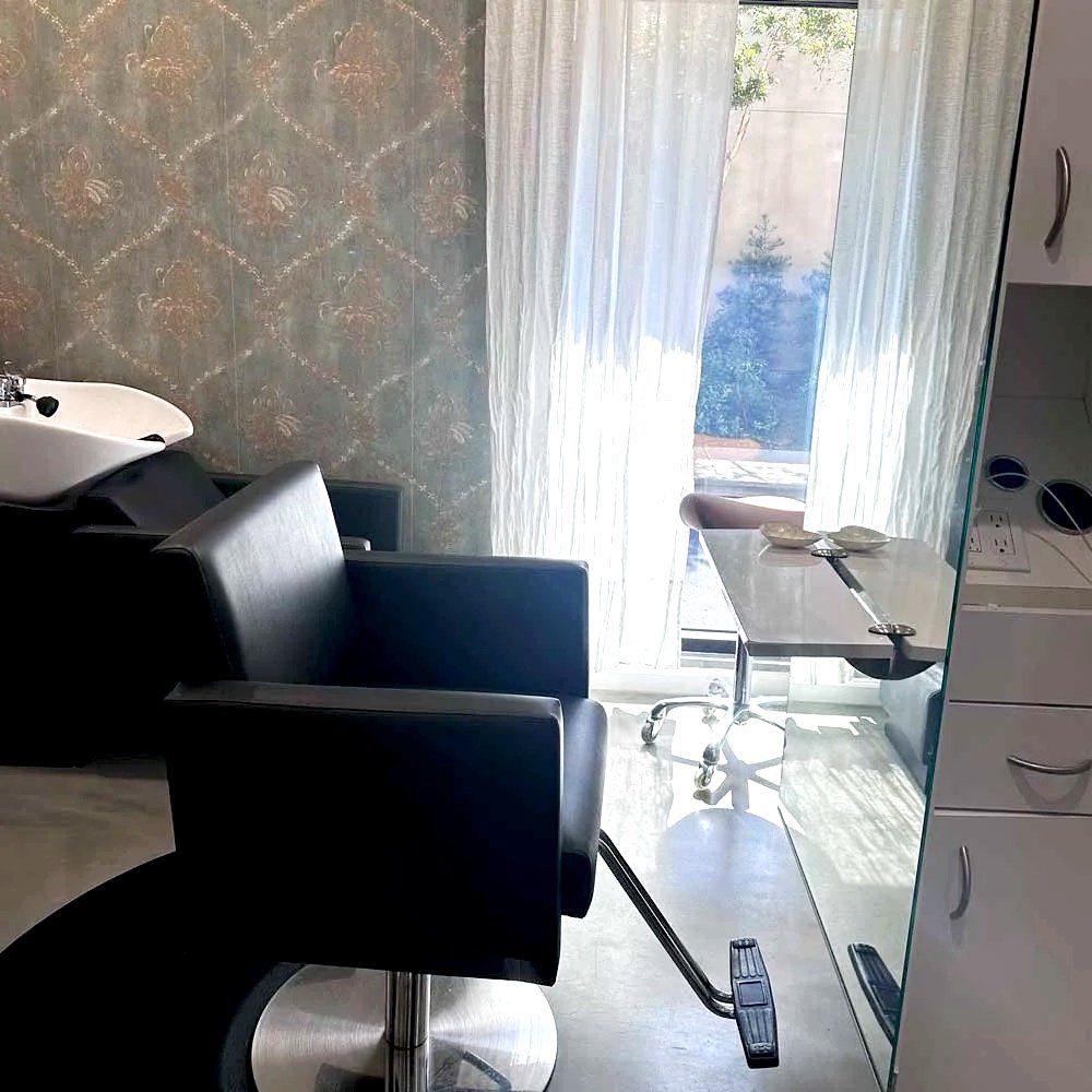 A hair salon or barber shop with a black styling chair in the foreground, a sink to the left, a small white table with a stool in the background, and sheer curtains covering a door or window with sunlight streaming through.