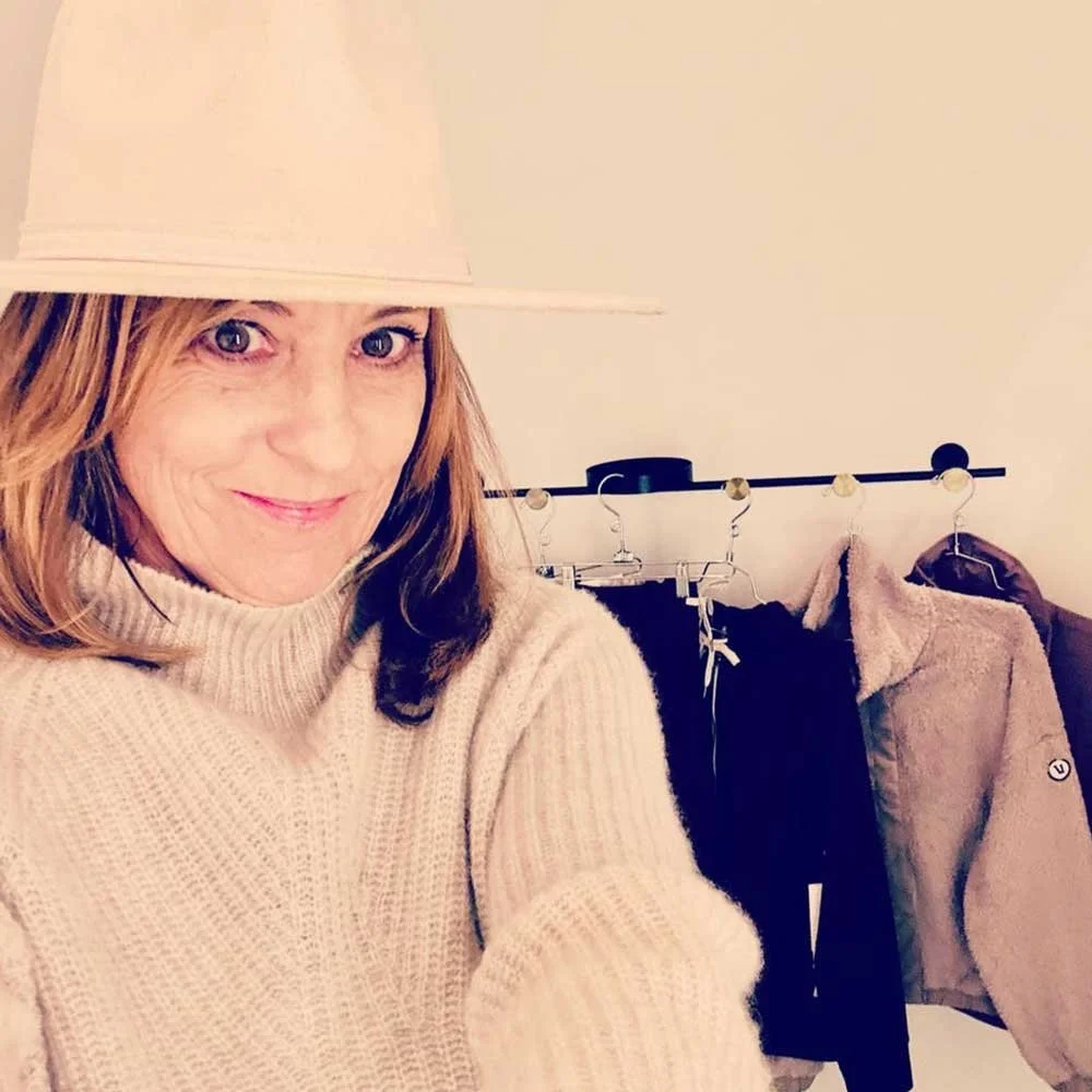A woman taking a selfie with a cream-colored turtleneck sweater and a beige hat, standing in front of a clothing rack with jackets and pants.