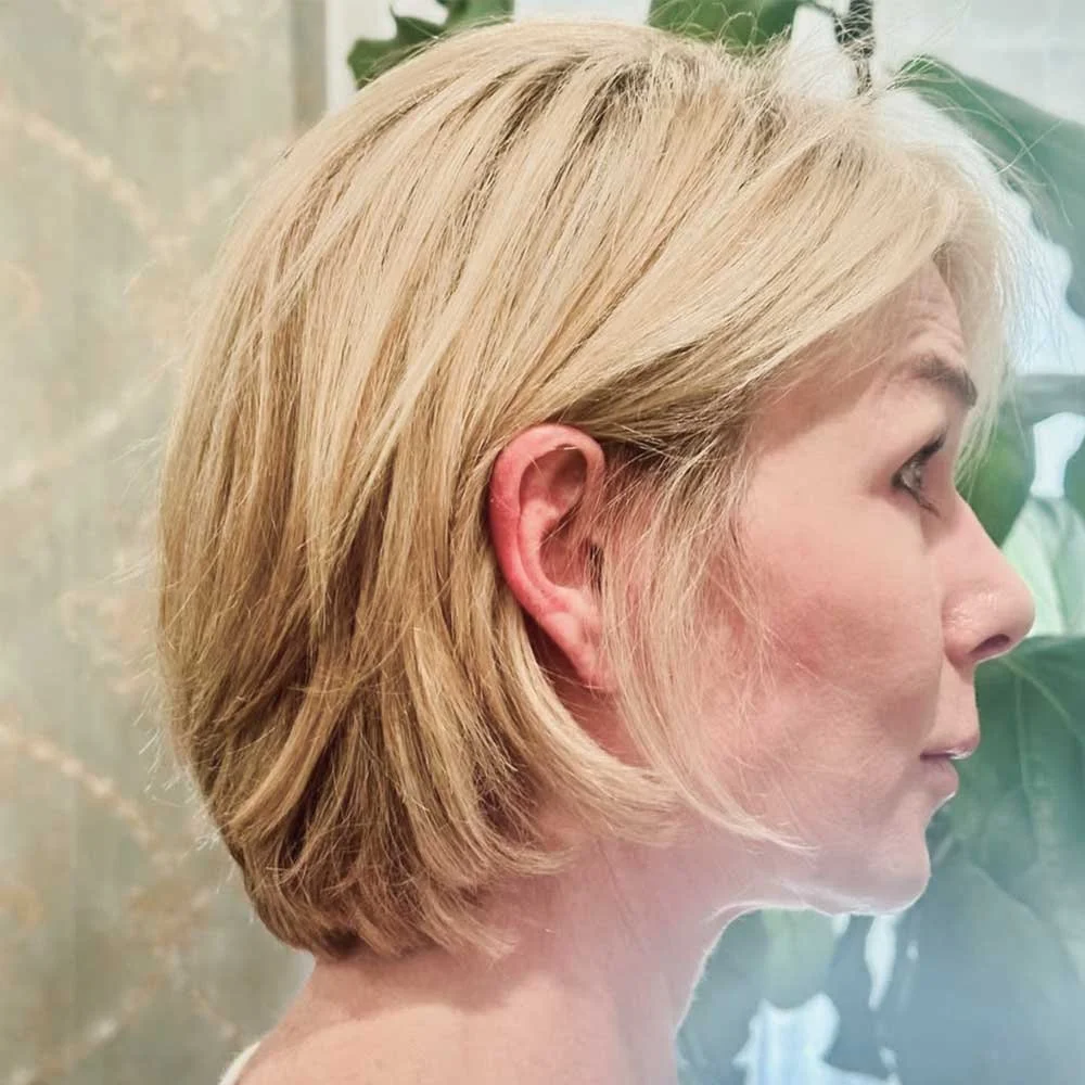 Side profile of a blonde woman with short hair, standing against a background of green leaves.