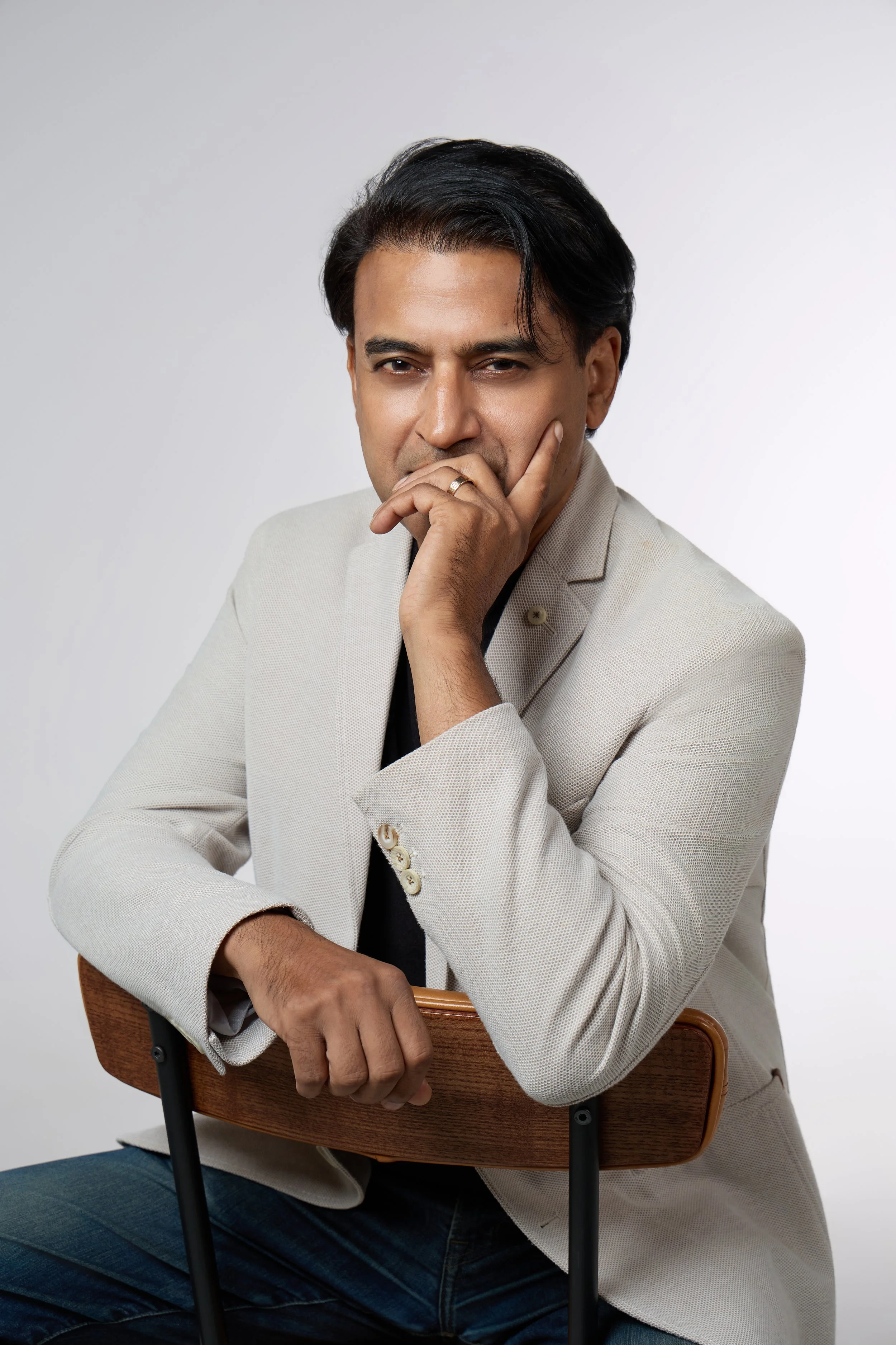 A man with dark hair, dressed in a beige blazer and black shirt, sitting on a chair with his arm resting on the back of a wooden chair. He is holding his chin with his hand, in a thoughtful pose, against a light gray background.