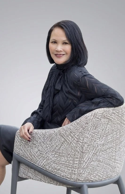 Portrait of a woman with shoulder-length black hair, wearing a dark patterned blouse, sitting on a light-colored armchair with geometric patterns, against a plain gray background.