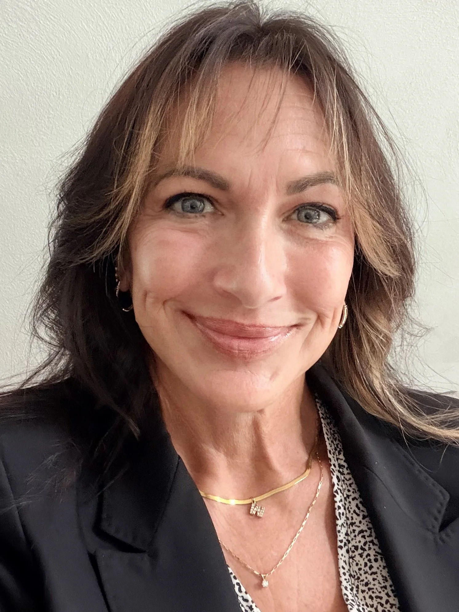 Smiling woman with shoulder-length brown hair, wearing a black blazer, leopard print top, gold jewelry including a necklace and earrings, standing against a neutral background.