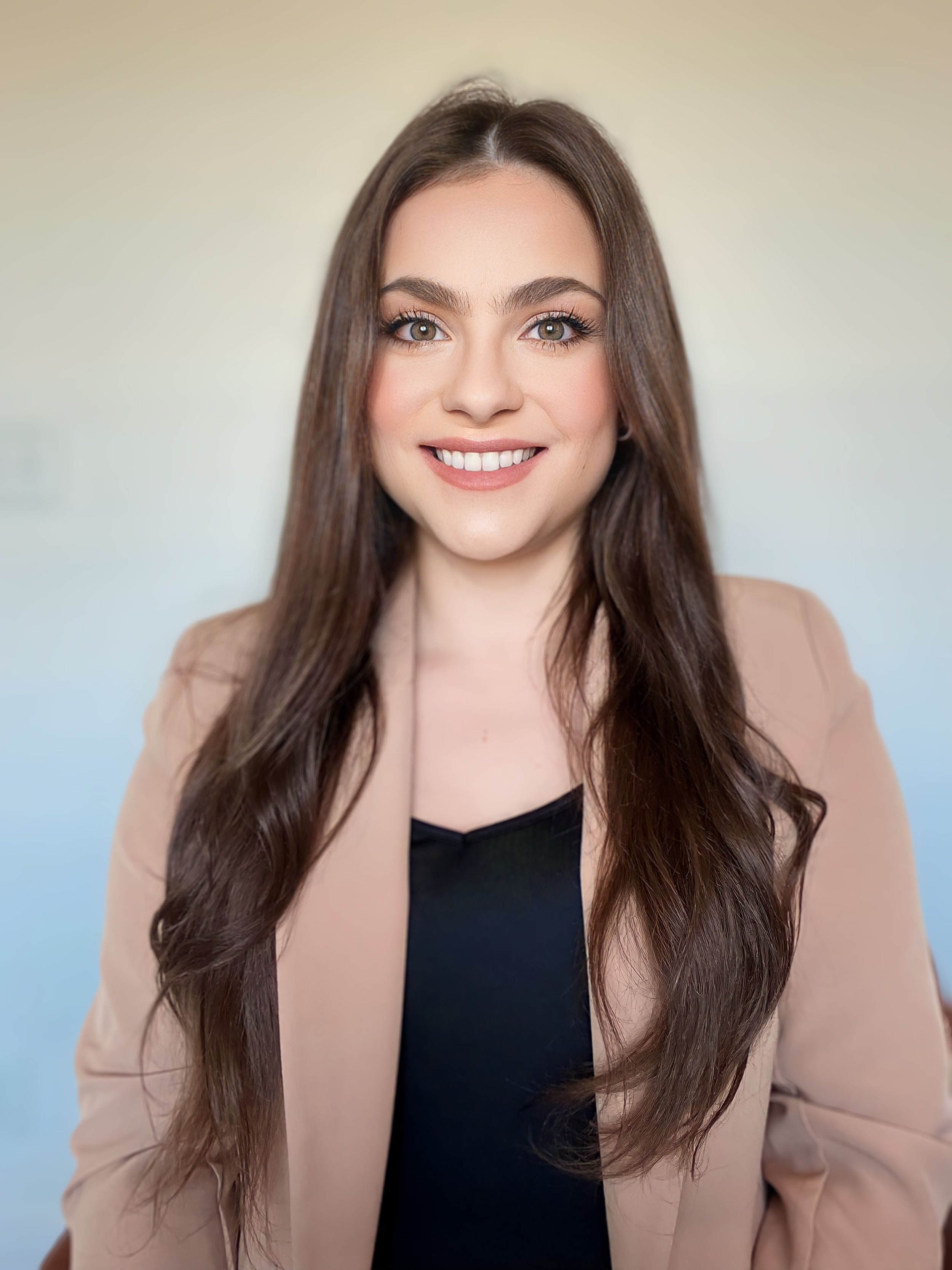A woman with long, wavy brown hair and gray eyes smiling at the camera, wearing a beige blazer over a black top, against a blurred light background.