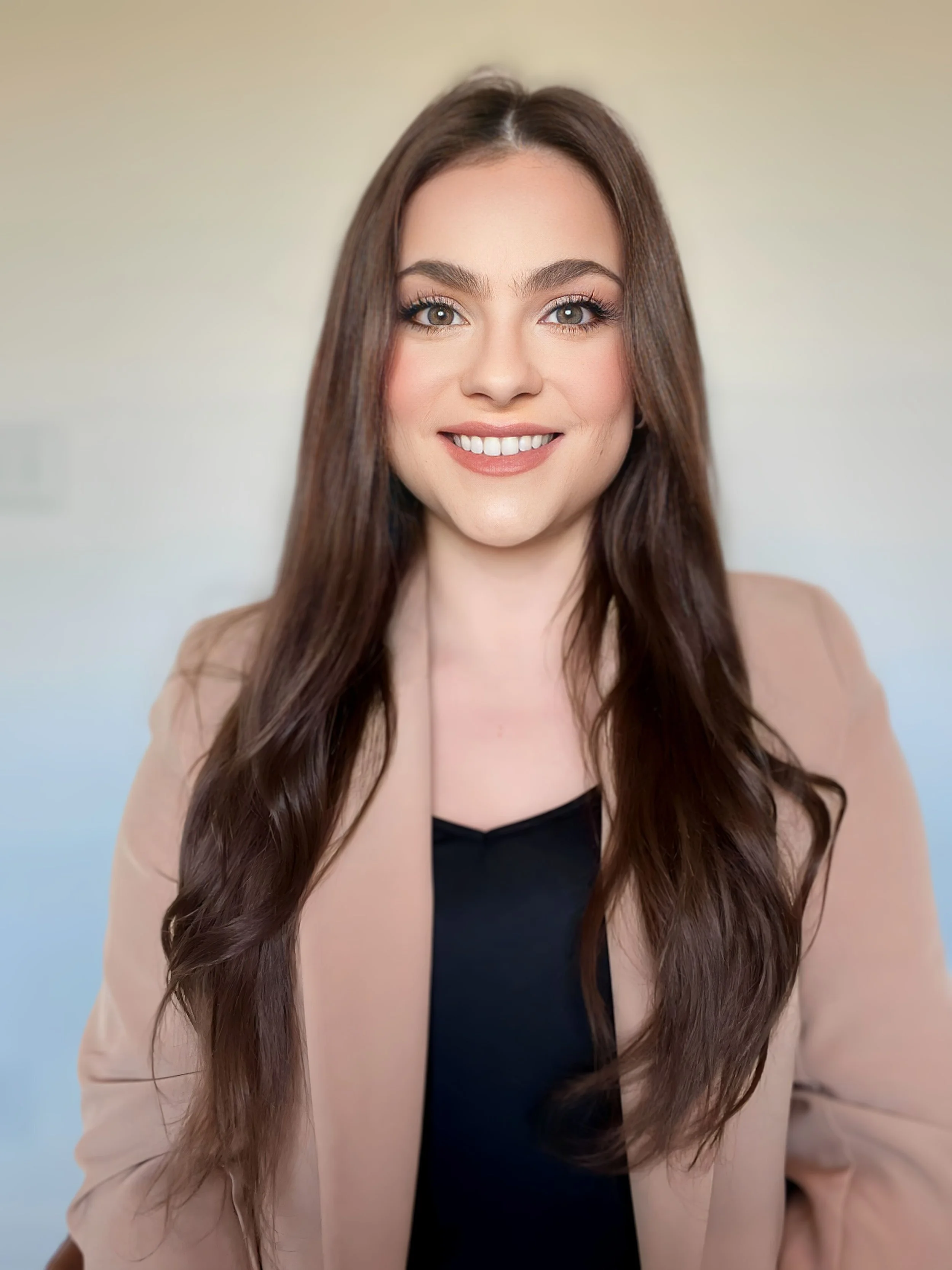 Portrait of a smiling young woman with long brown hair, wearing a beige blazer over a black top, standing in front of a plain light background.