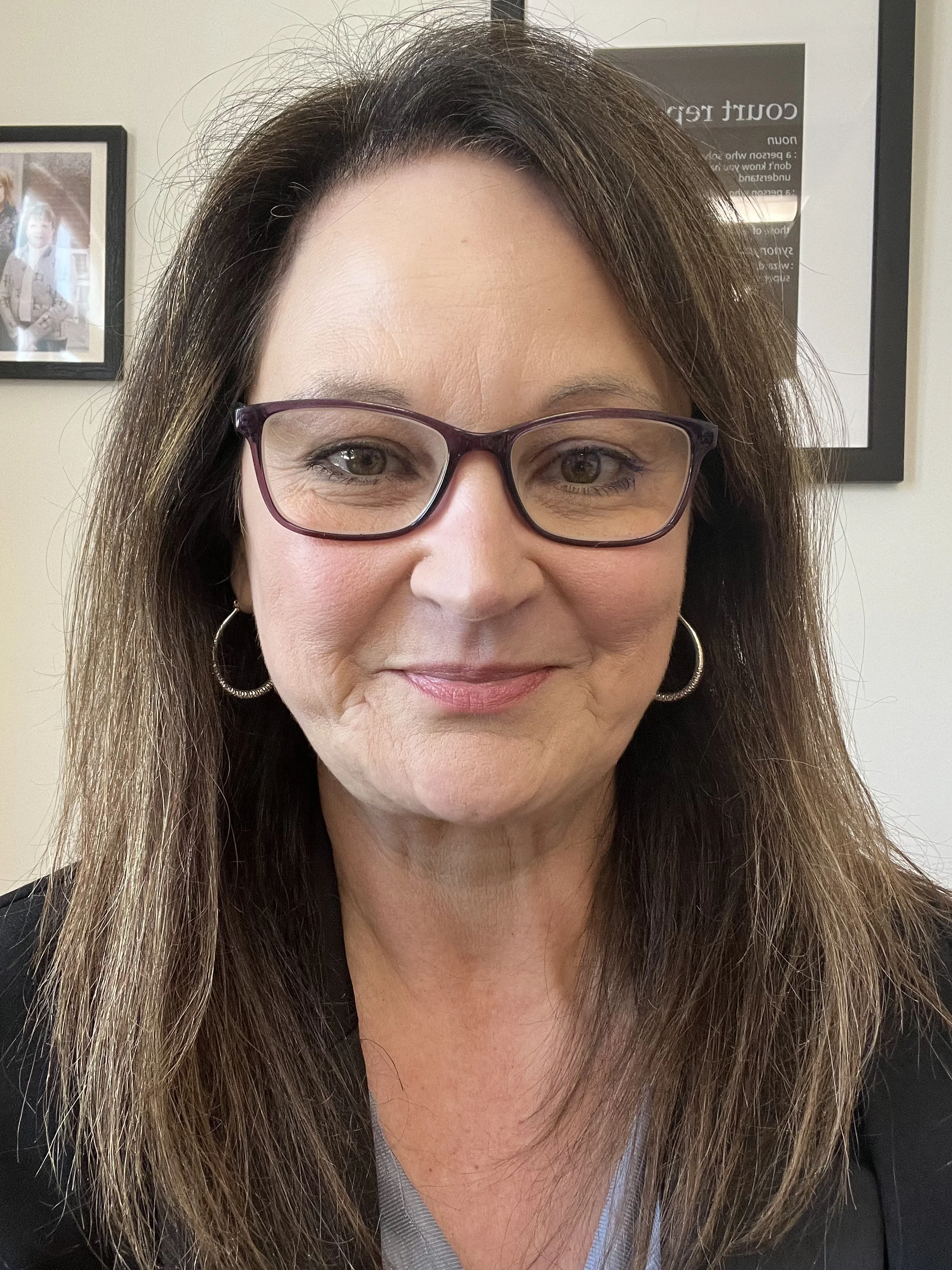 A close-up photo of a woman with long brown hair, wearing glasses with purple frames, silver hoop earrings, and a black jacket. Behind her, there are framed pictures and a sign on the wall.