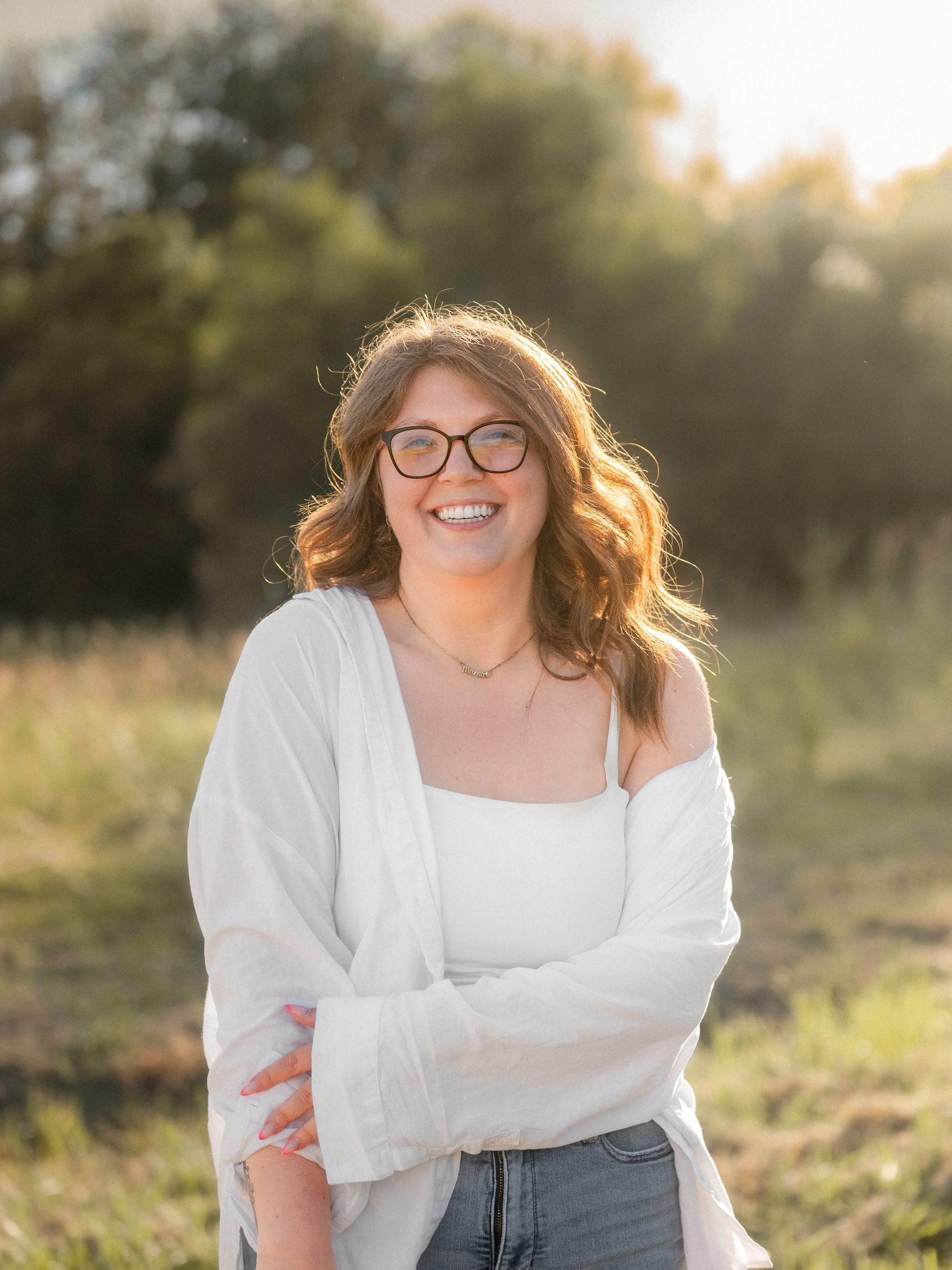 A smiling young woman with curly brown hair and glasses standing outdoors in a grassy area with trees in the background, illuminated by warm sunlight.