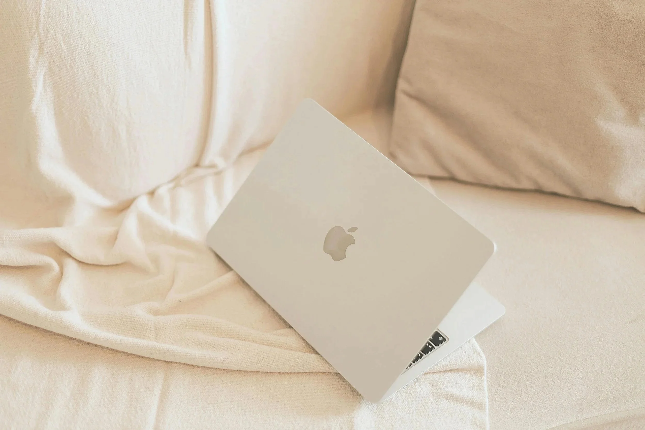A closed silver Apple MacBook laptop on a cream-colored satin or silk bedsheet with a beige pillow in the background.