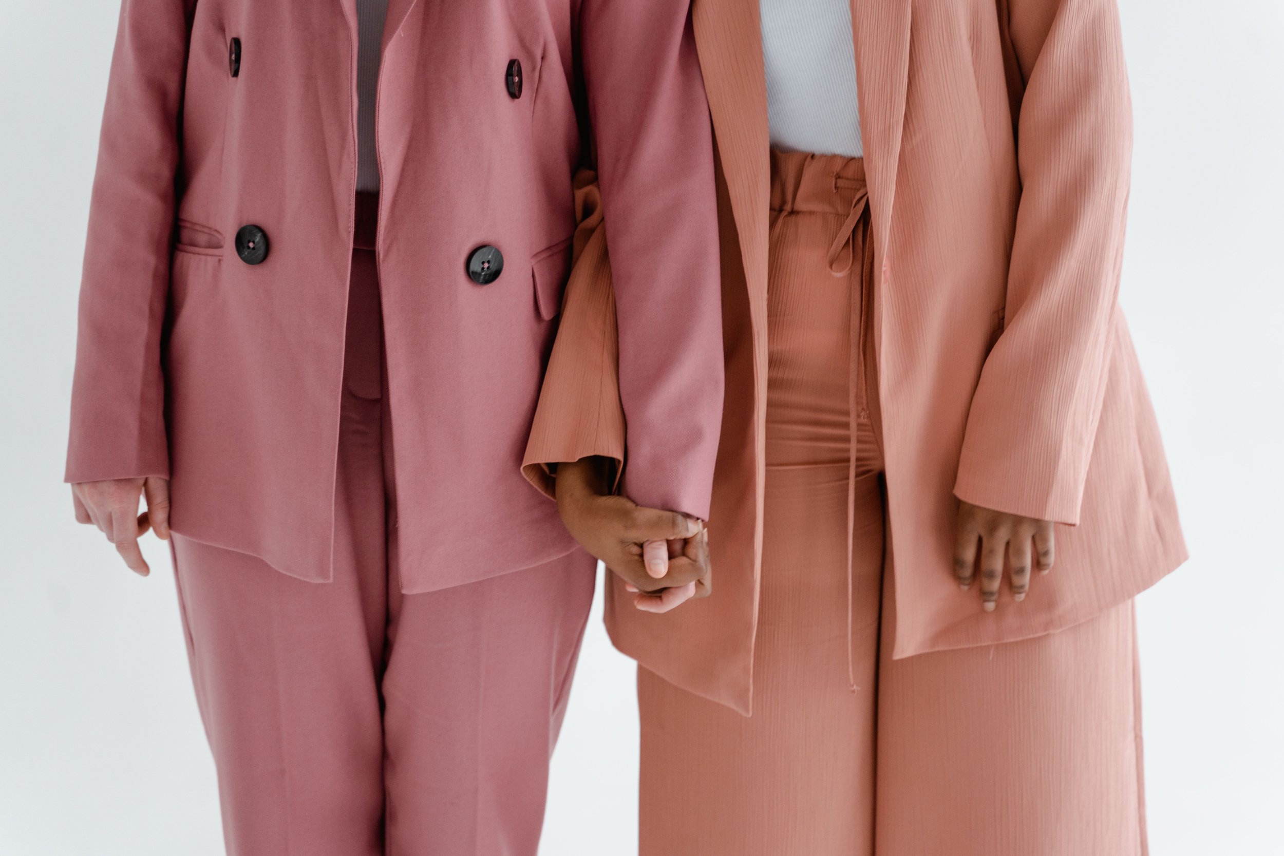 Two people holding hands dressed in pink and peach-colored suits, standing against a white background.