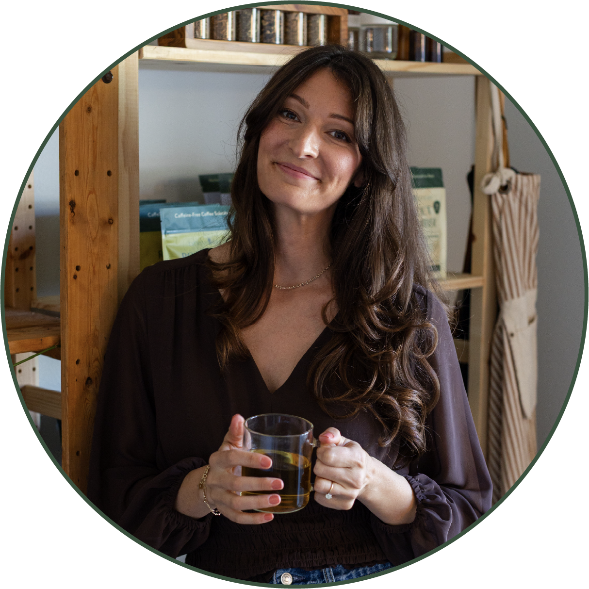 A woman with long brown hair holds a glass of tea, smiling at the camera in a cozy setting with wooden shelves and coffee bags in the background.
