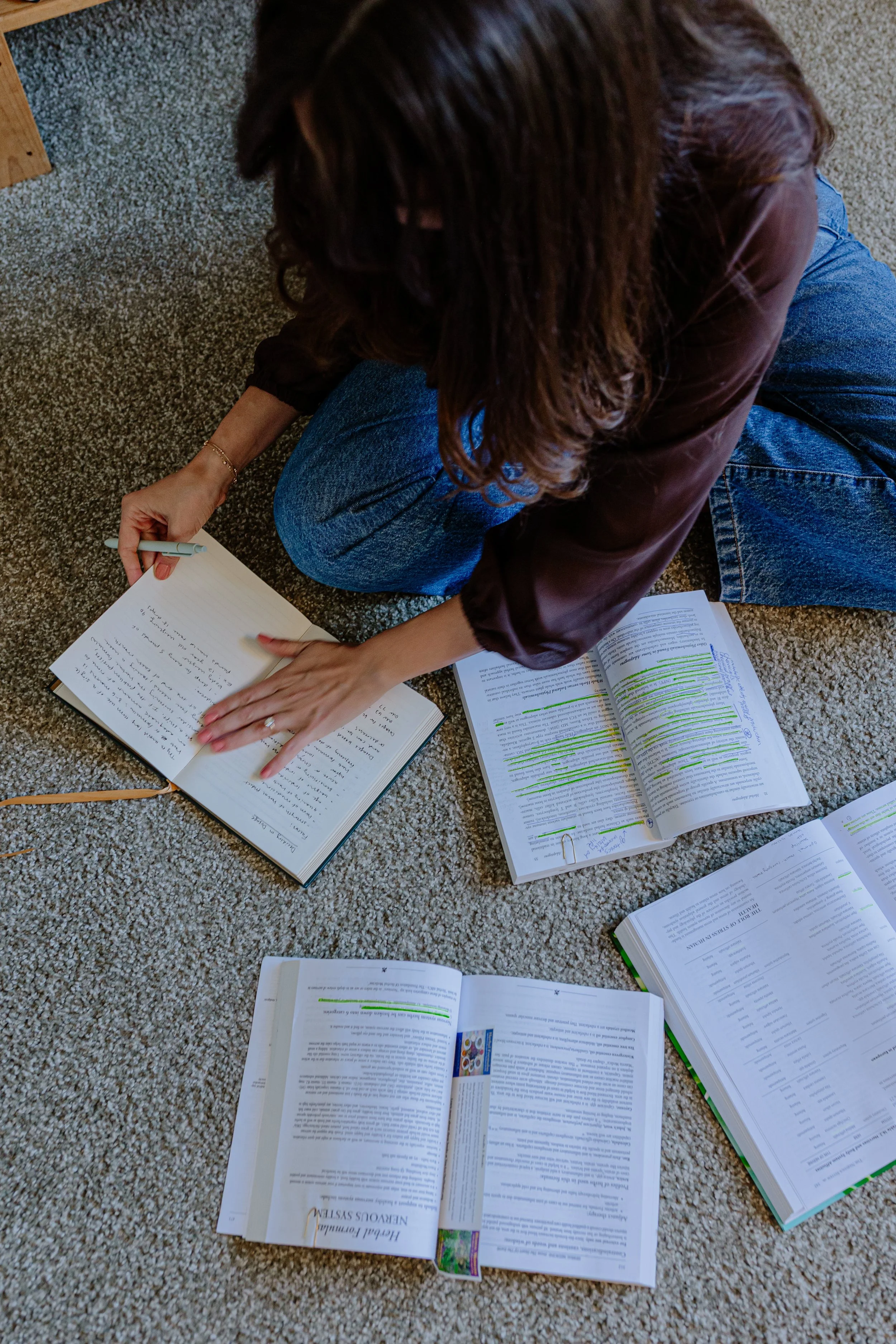 Person sitting on carpeted floor with multiple open books and notes, writing in one book with a pen.