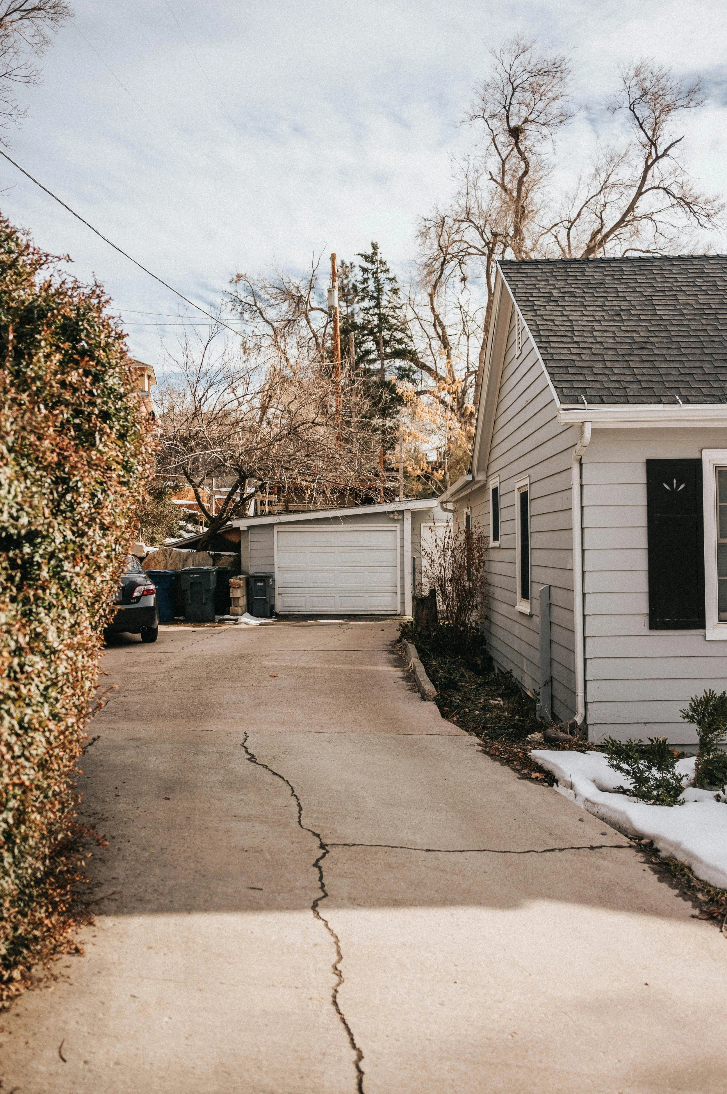 A driveway with a crack running through it, next to a white house with black shutters, some snow, and a garage at the end in Falls Church, VA