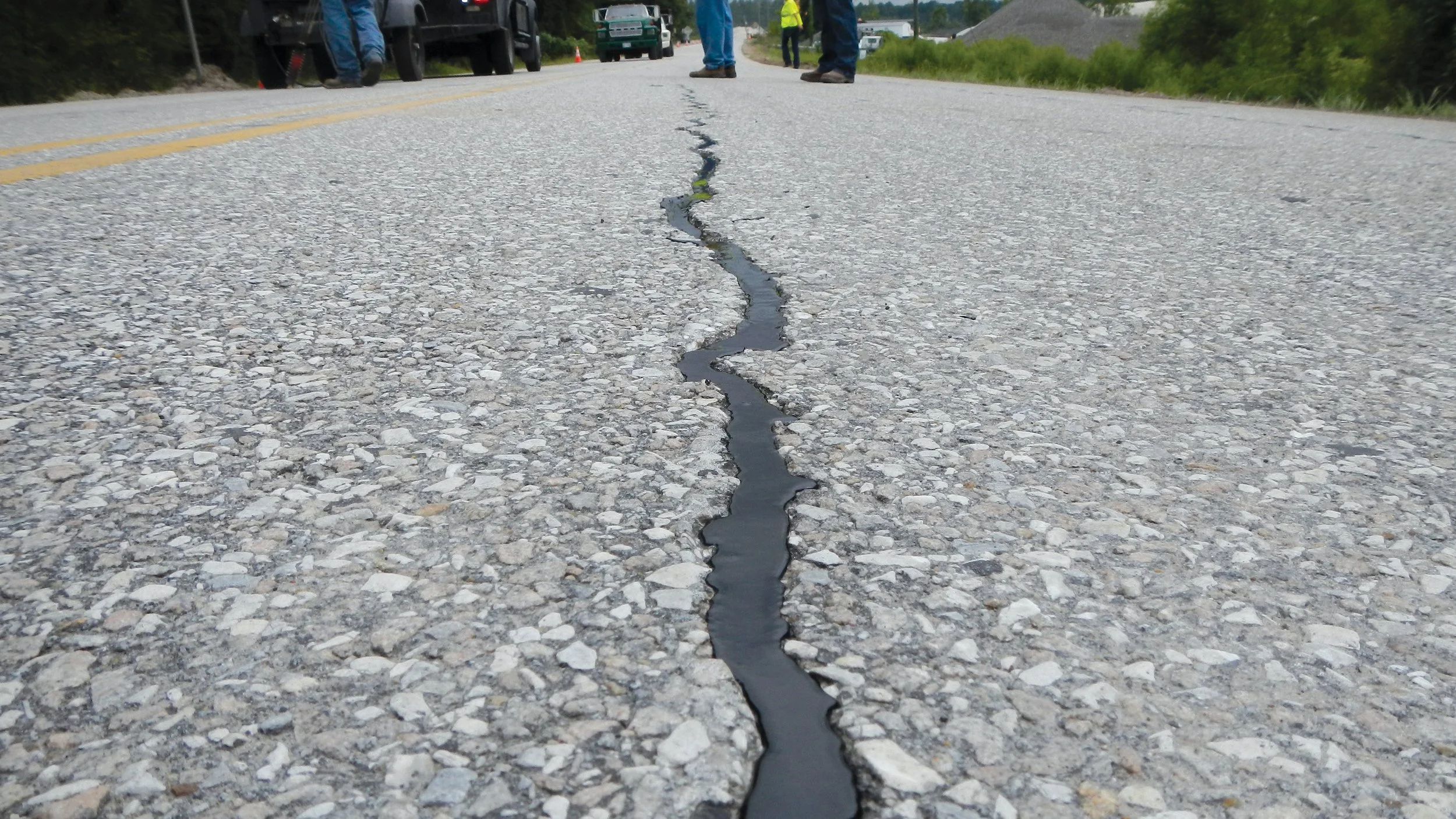 Close-up of a cracked asphalt road with repair material along the crack, and several people and vehicles in the background.