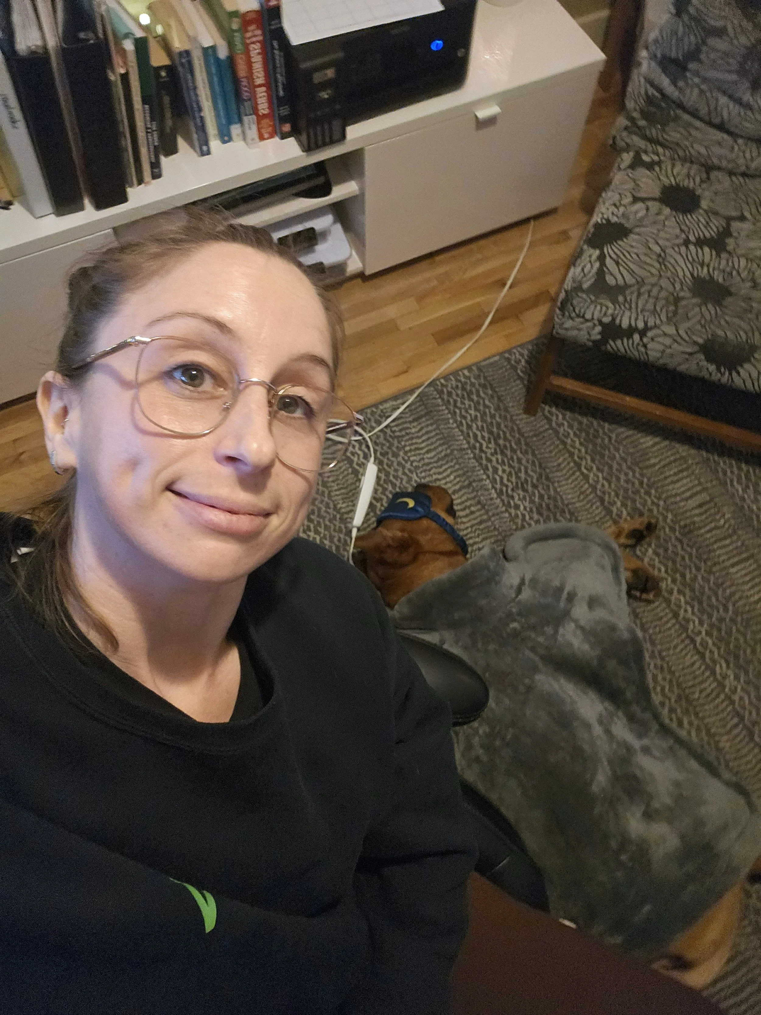 A woman with glasses smiles in a selfie, sitting on the floor of a cozy living room with a dog resting nearby