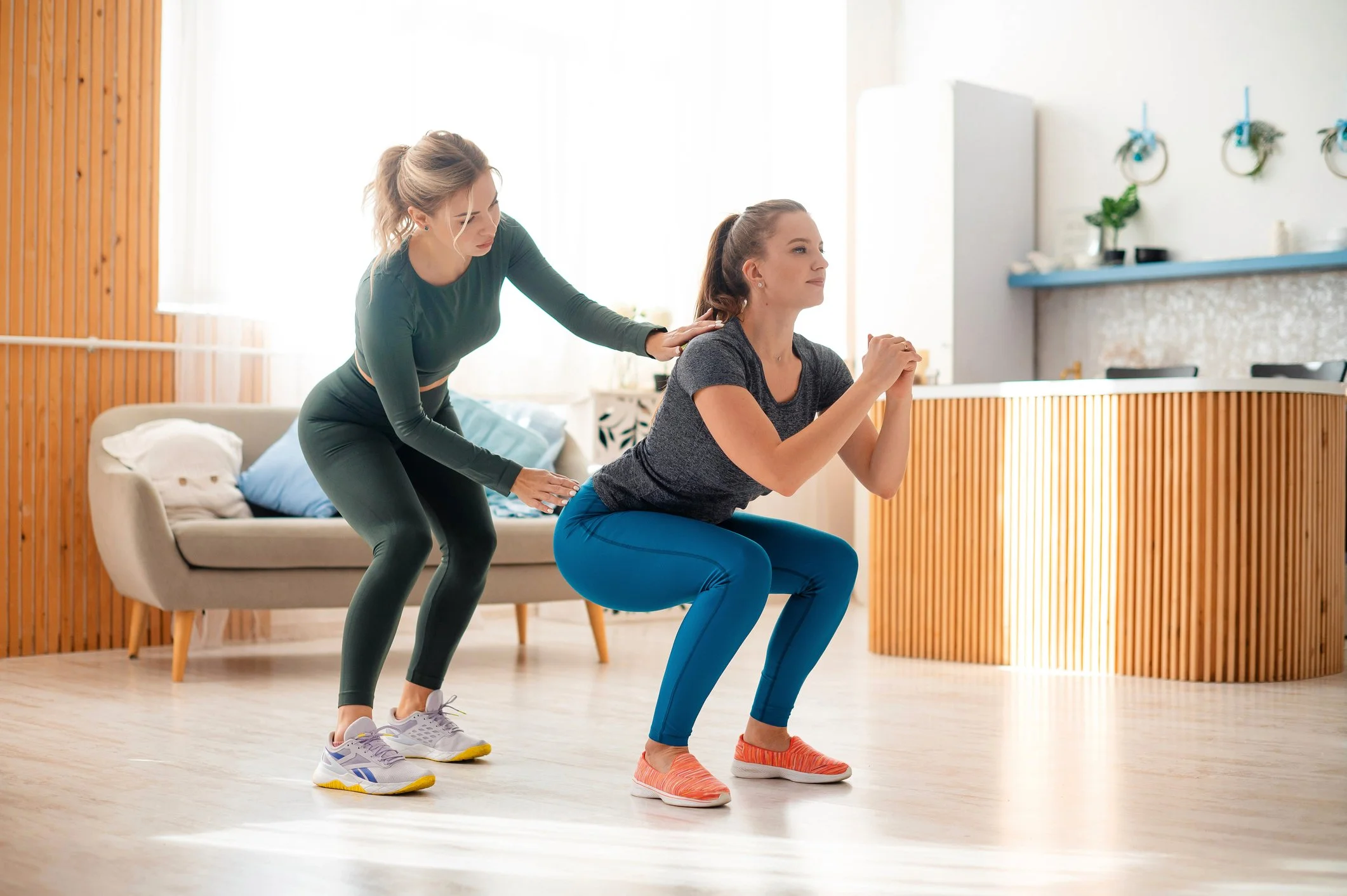 Two women exercising indoors; one is leading a squat, the other is assisting, in a bright room with wooden and white decor.