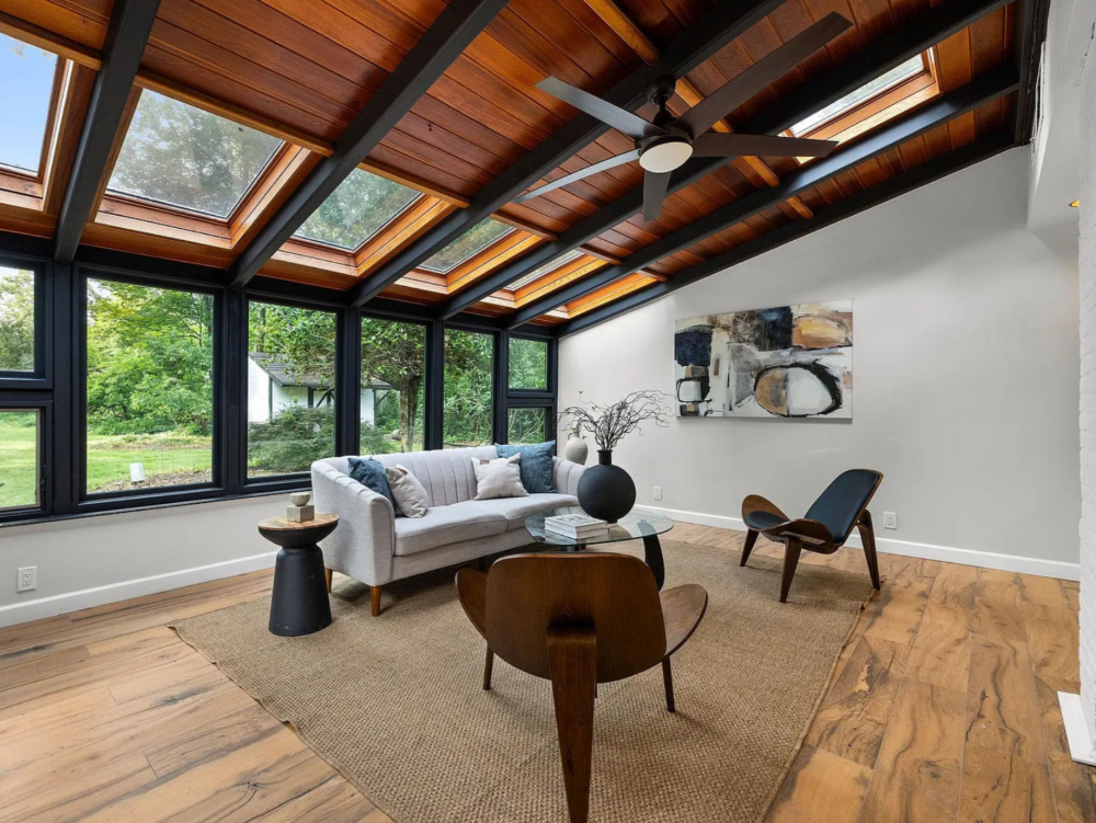 Living room with large windows, wooden ceiling with skylights, ceiling fan, white sofa with decorative pillows, wooden chairs, art piece on white wall, glass coffee table with books and vase, area rug, wooden floor, and greenery outside.