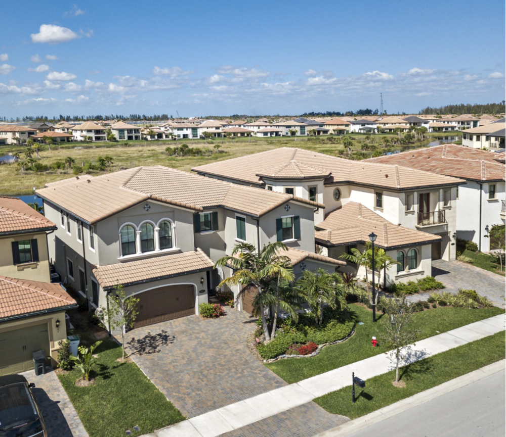 Aerial view of a suburban neighborhood with Mediterranean-style houses, green lawns, and palm trees, under a partly cloudy sky.