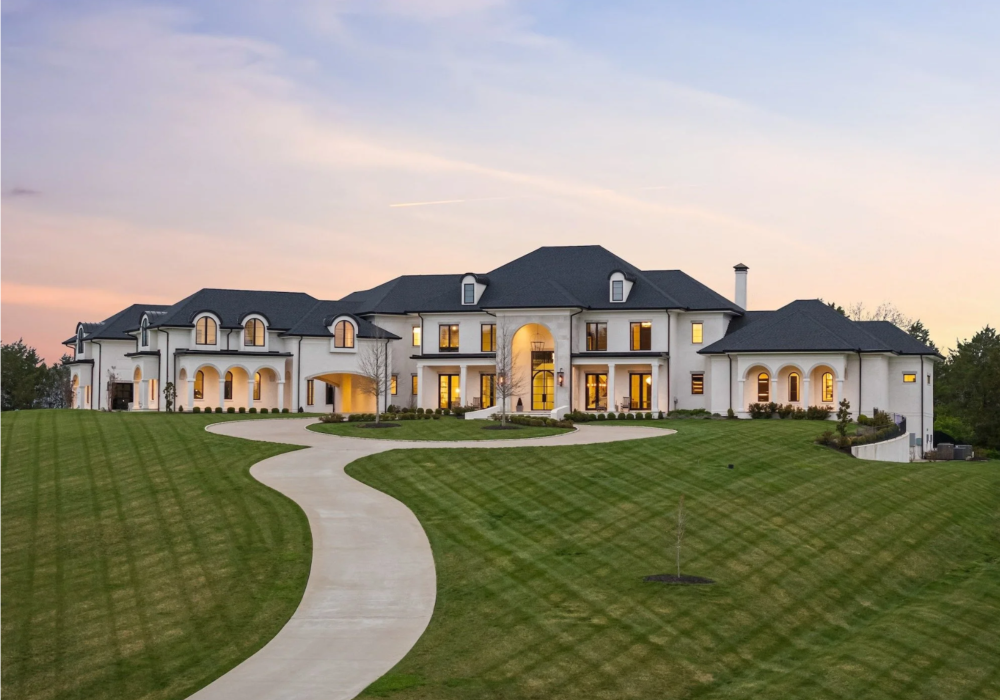 Large luxurious house with a curved driveway, well-manicured lawn, and multiple windows, at sunset.