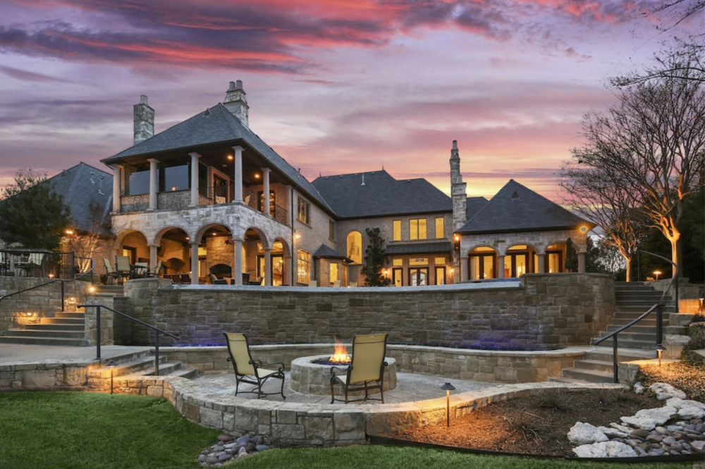 Luxury house with stone facade and multiple chimneys, illuminated at dusk, with a backyard featuring a fireplace and outdoor seating area, landscaped with grass, rocks, and trees.