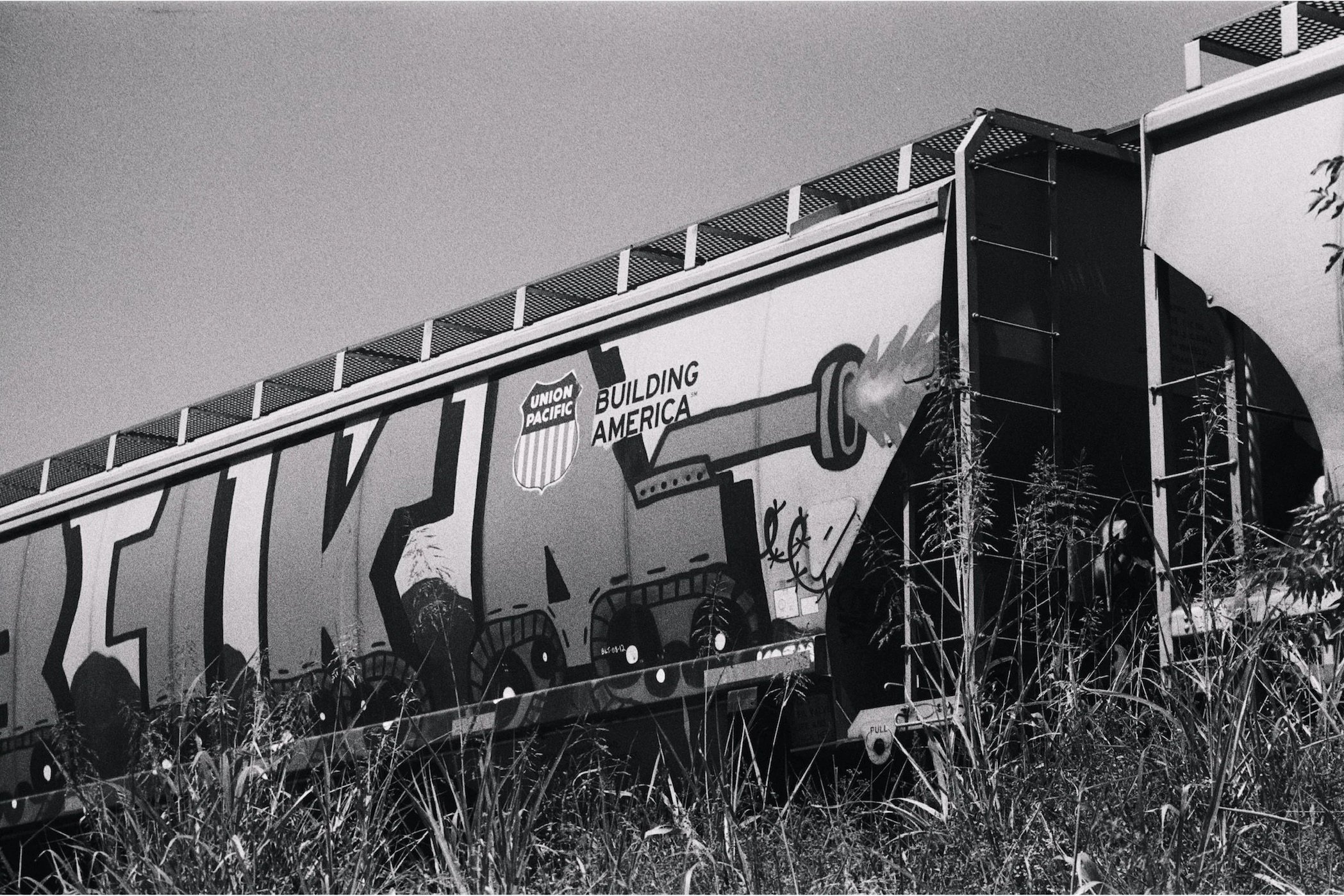 Black and white photo of a train car with Union Pacific logo and a mural of a torch, mountains, and trees, surrounded by tall grass.