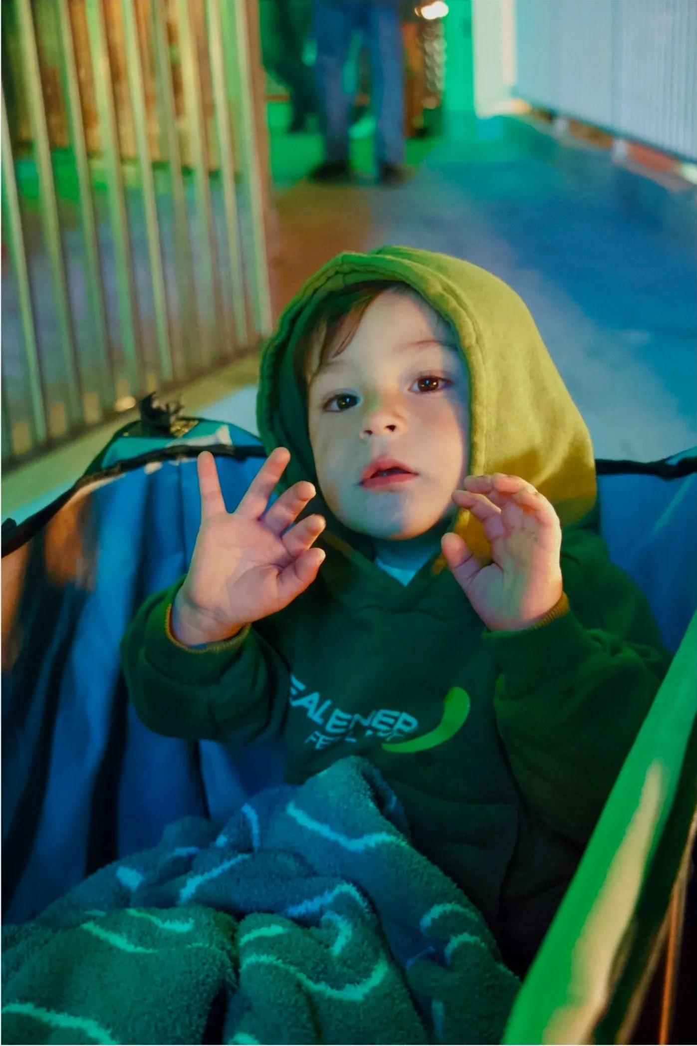 A young boy with a green hooded sweatshirt sitting in a stroller, waving at the camera, covered with a blanket in an outdoor setting at night with colorful lighting.