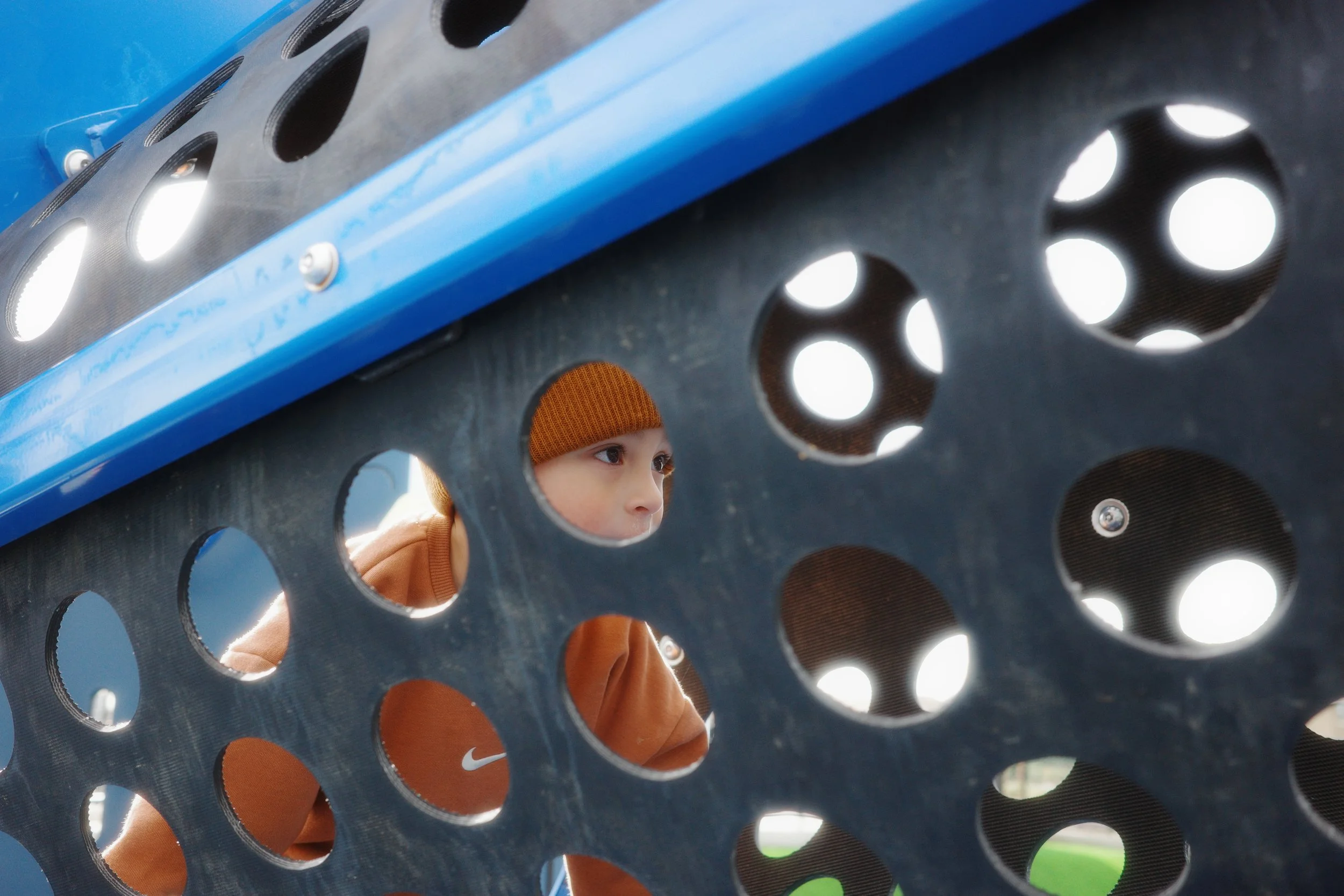 A young boy wearing an orange beanie and brown jacket looks through a large hole in a metal play structure at a playground.