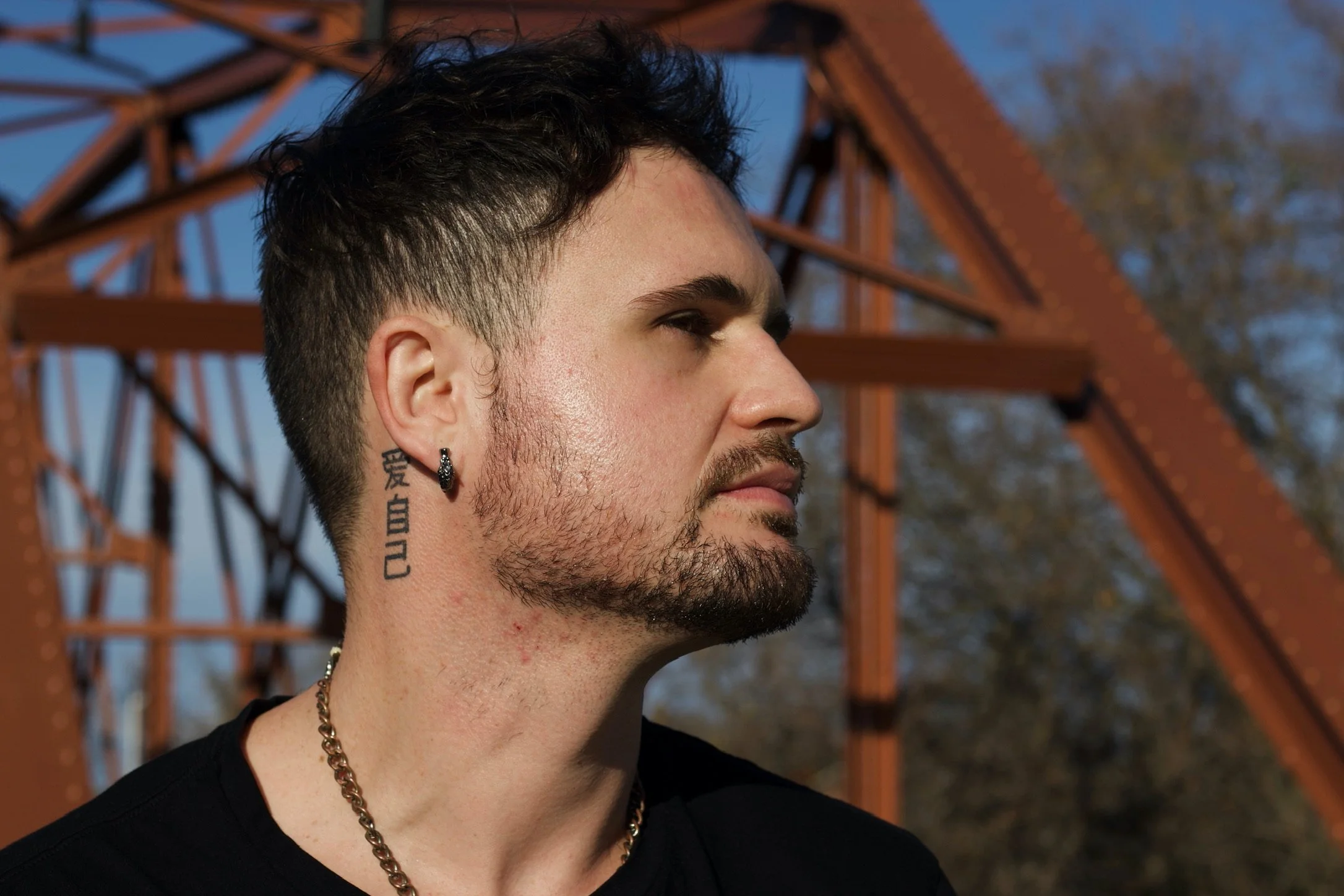 A young man with dark, styled hair and a beard, wearing a black shirt, a silver chain necklace, and a black earring, stands outdoors against a rusty red metal bridge with a sunset sky and trees in the background.