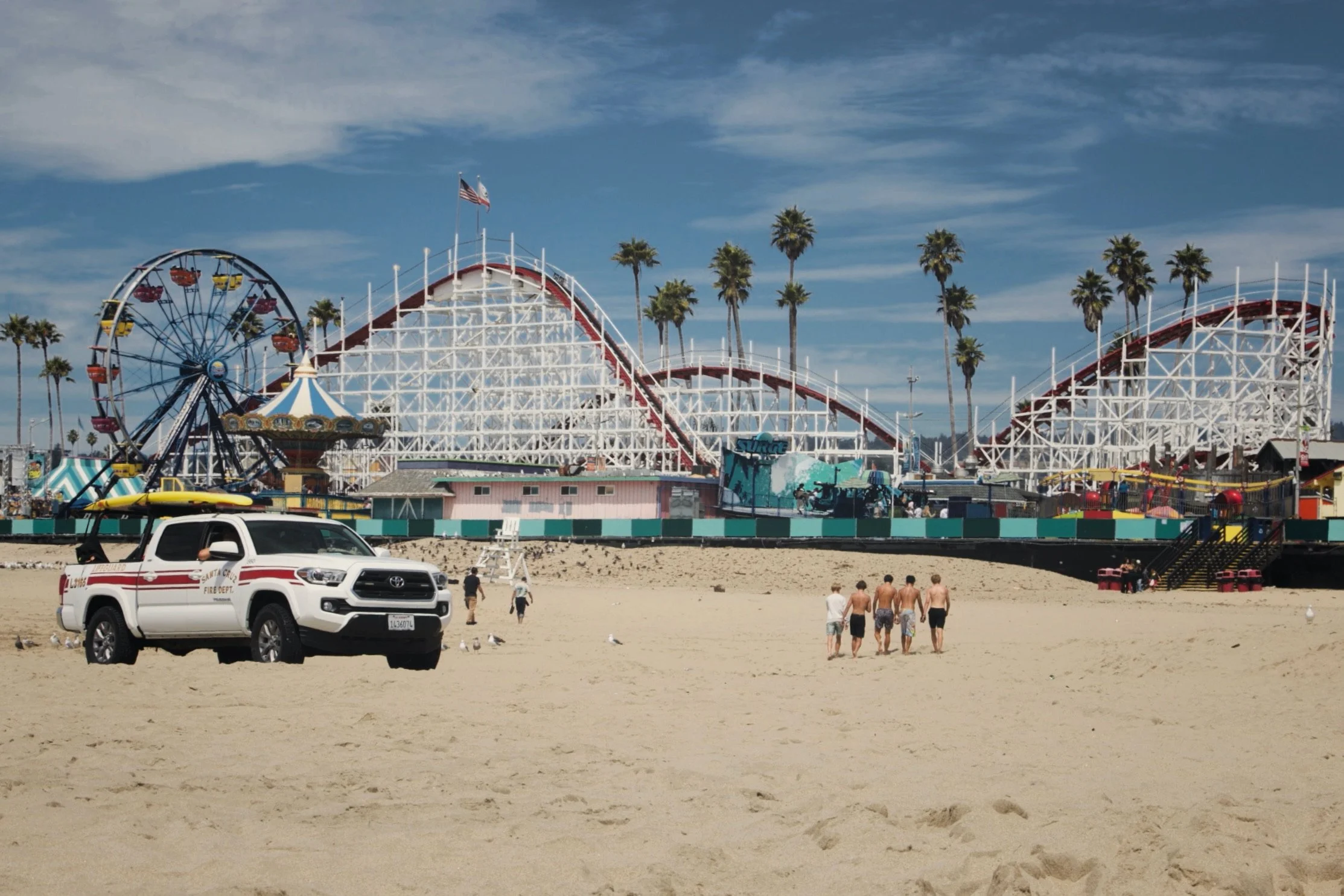 Beach scene with an amusement park featuring a roller coaster, a Ferris wheel, and a carousel, with a fire department truck and four people walking on the sand, and palm trees under a partly cloudy sky.