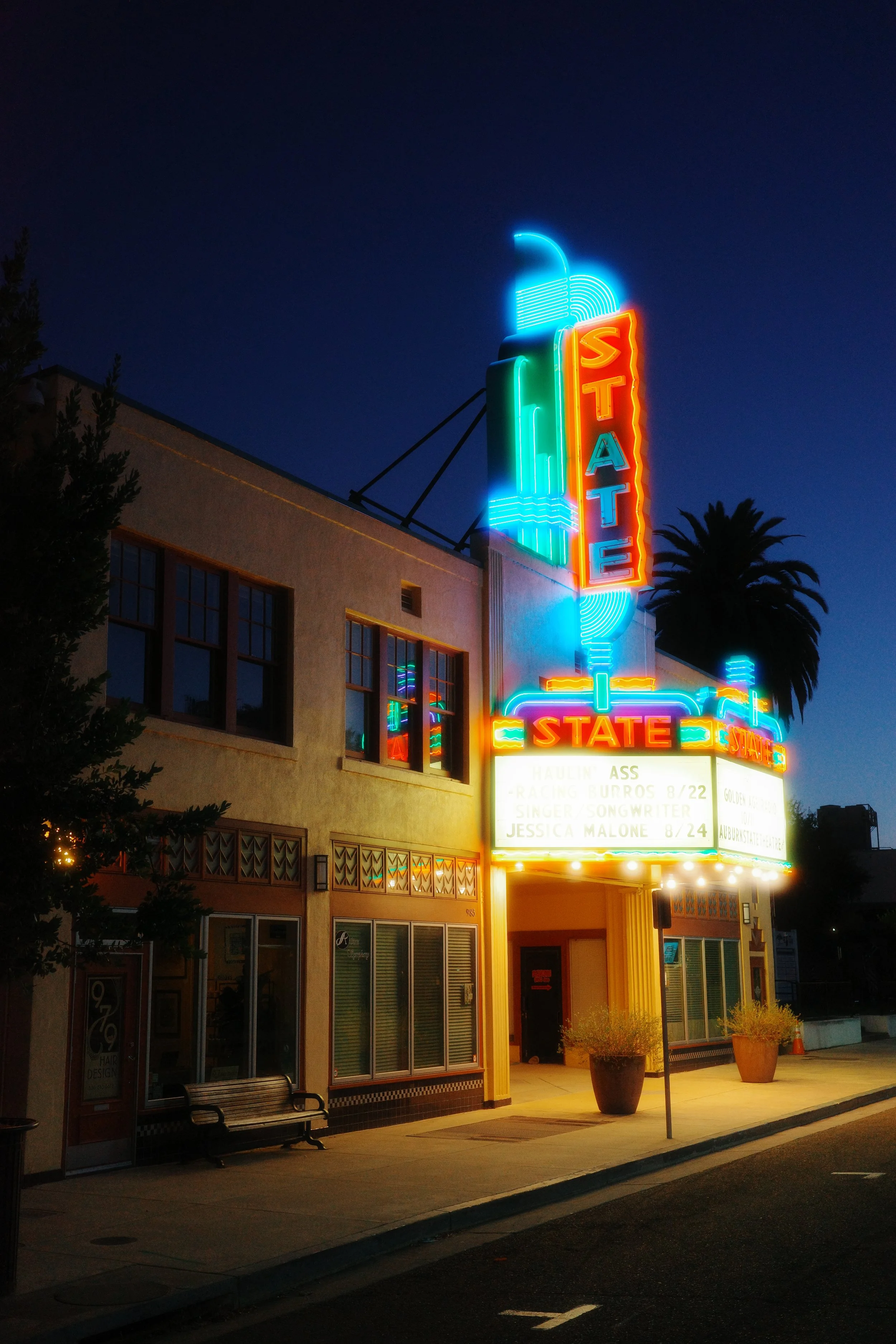 Night view of a vintage theater building with bright neon sign that reads 'STATE', with additional neon lights and a marquee advertising upcoming performances.