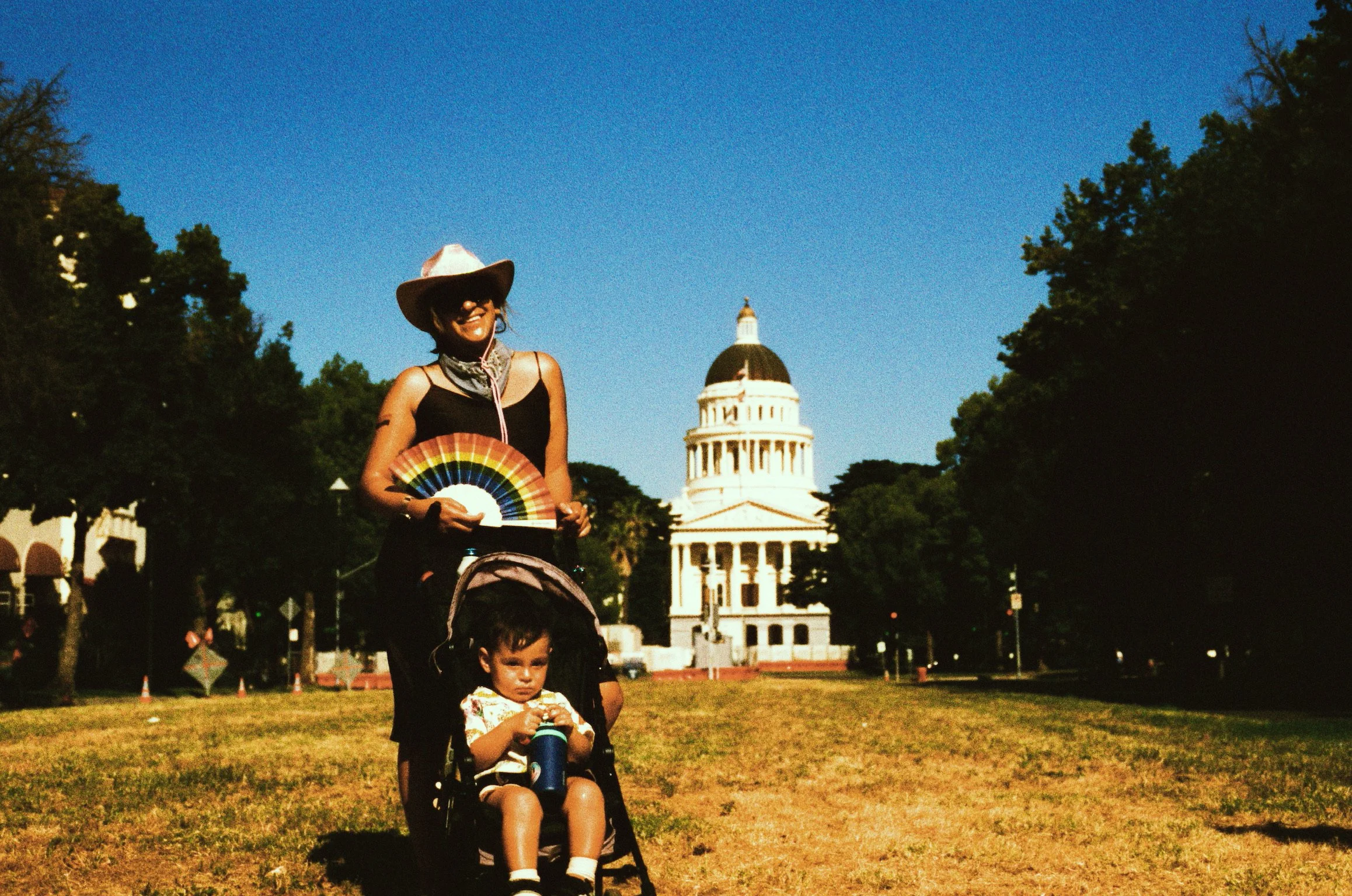 A woman with a large sun hat and sunglasses pushing a stroller with a young boy holding a cup, on a grassy area with the Capitol building in the background under a clear blue sky.