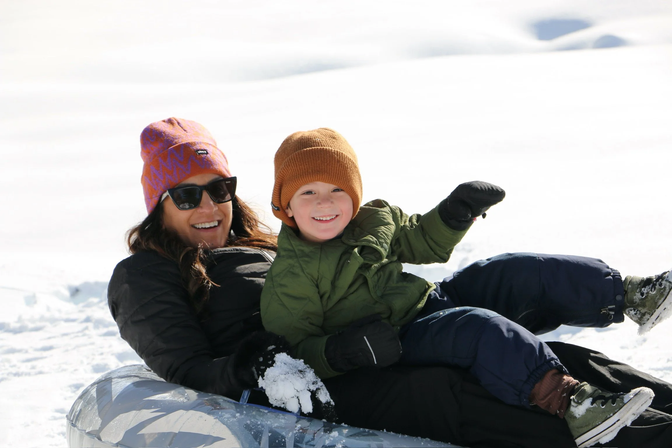 A woman and a young boy enjoying snow tubing in a snowy outdoor setting. They are dressed in winter clothing, smiling and having fun.