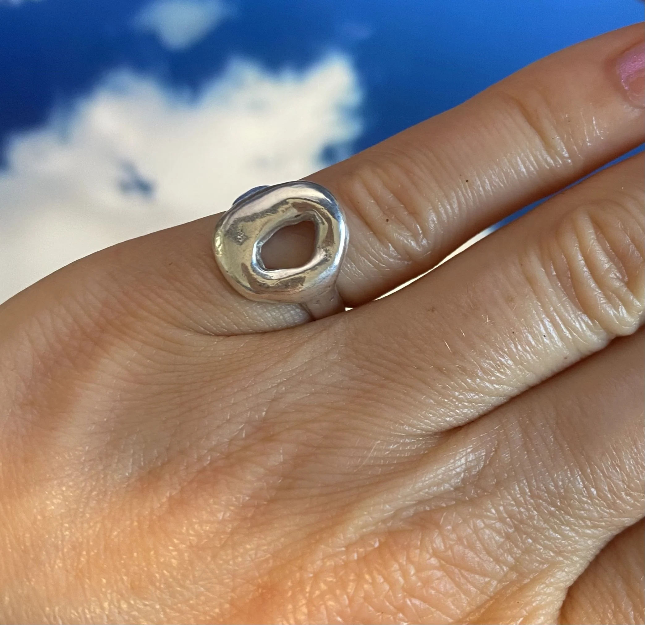 Ring aus Solid Silver auf dem kleinen Finger. Er glänzt und sieht aus wie ein kleiner silberner Donut. Im Hintergrund-Wolken. Der Ring ist das Ergebnis eines Silver Clay Workshops von Studio Jupiter in Hamburg.