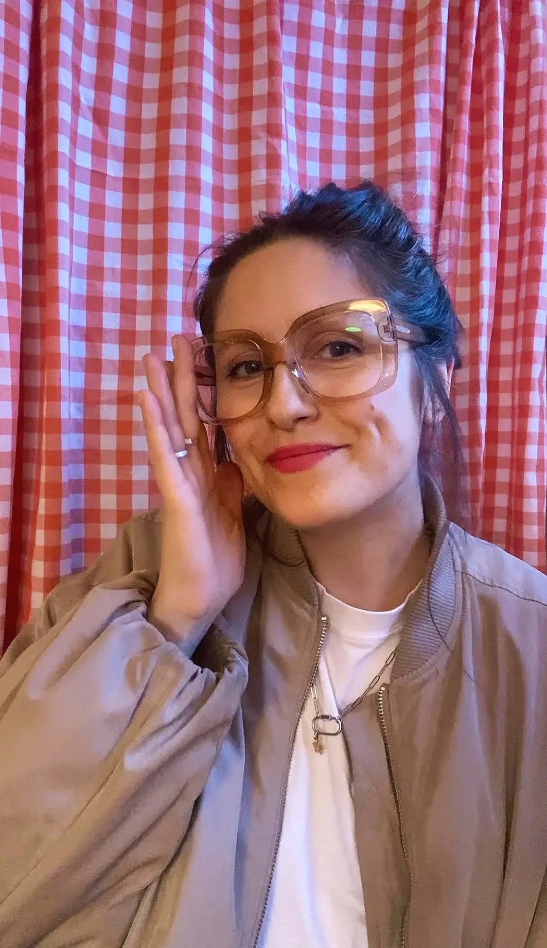 A woman , Linna Grage, with dark hair, wearing large pink glasses, red lipstick, a beige jacket, and a white shirt, smiling, with a red and white checkered curtain in the background.