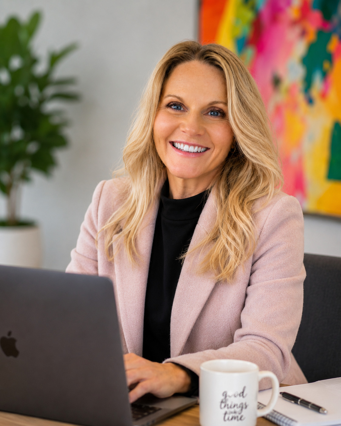 A woman with blonde hair smiling while sitting at a desk with a laptop, coffee mug, notepad, and pen, with a colorful abstract painting and a potted plant in the background.