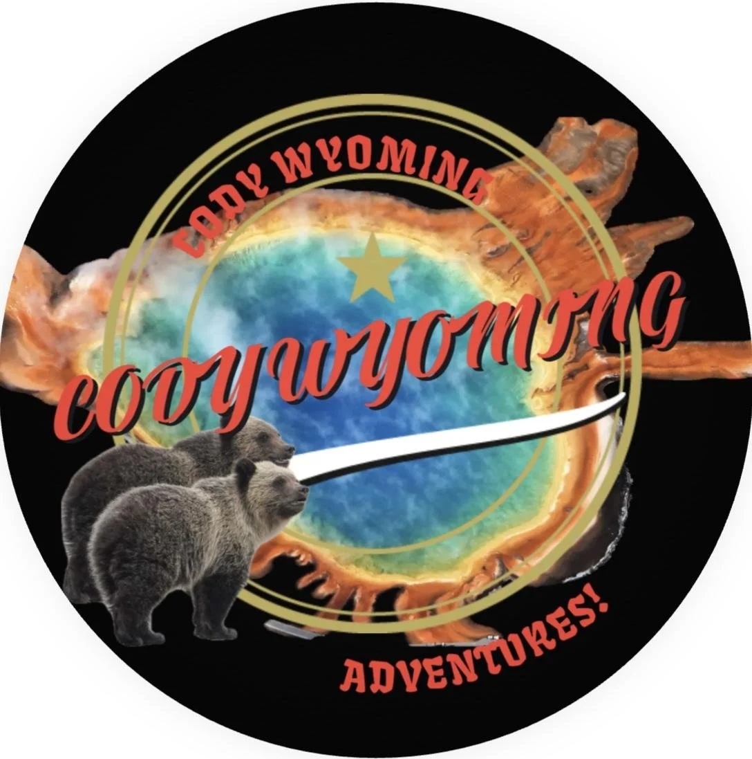 A logo for Cody Wyoming Adventures featuring a colorful earth globe at the center, with a trail of flames behind it. The text "Cody Wyoming" is curved at the top, and "Adventures!" at the bottom in red font. A bear is pictured in the bottom left corner.