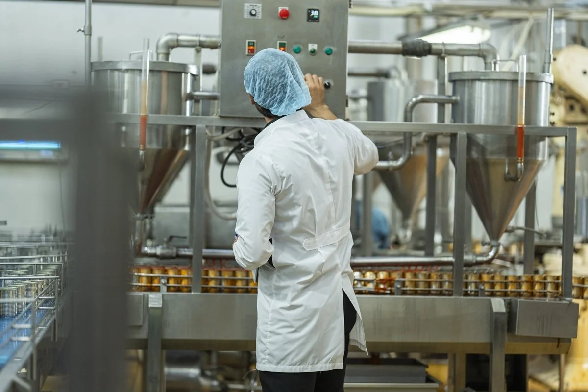 A worker in a white coat and blue hairnet operating industrial machinery in a manufacturing facility.