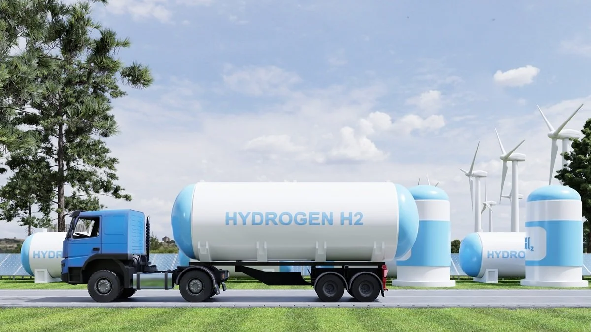 A blue truck carrying a large white tank labeled " hydrogen H2," driving past wind turbines and solar panels in a landscape with green grass, trees, and a partly cloudy sky.