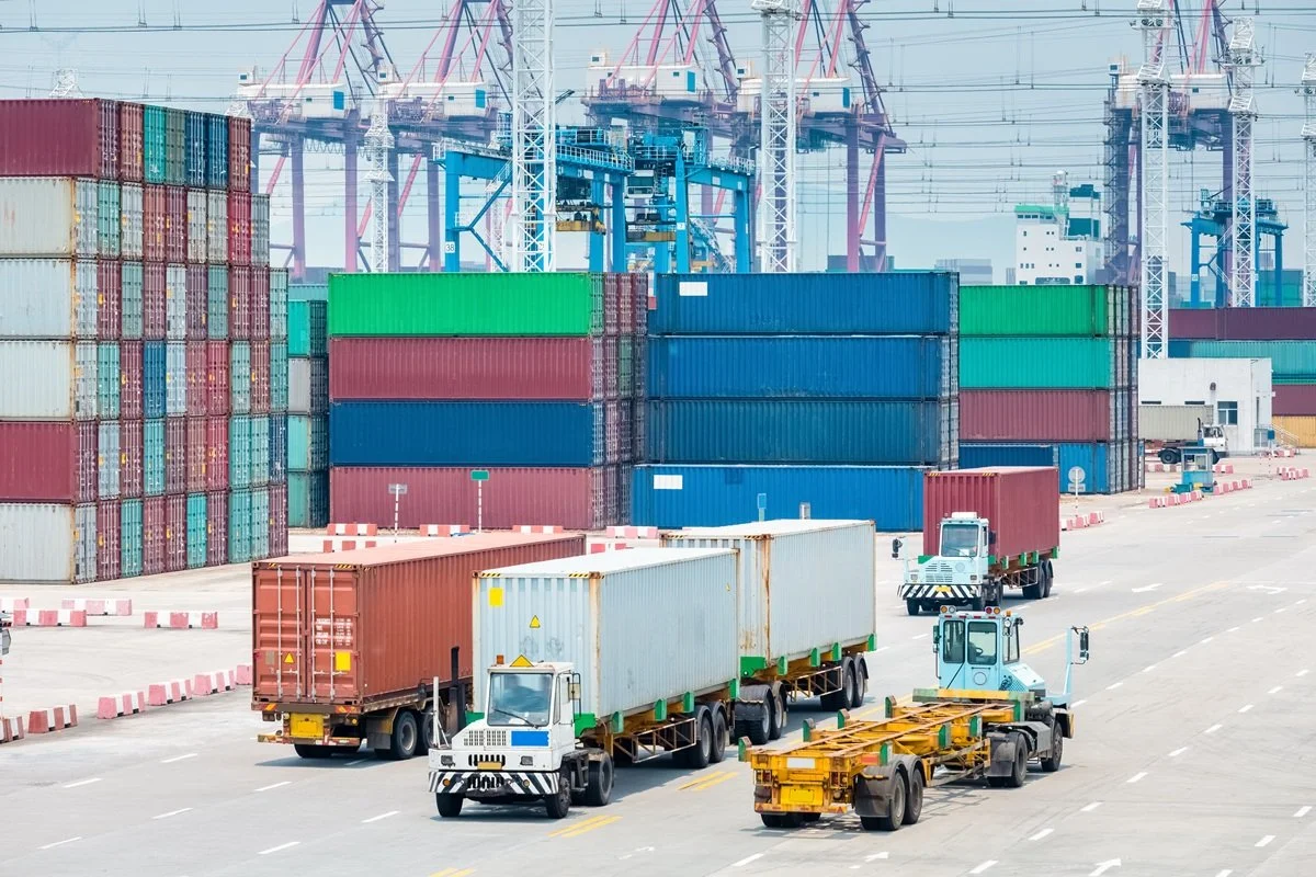 Containers stacked at a port with trucks and cranes in the background.