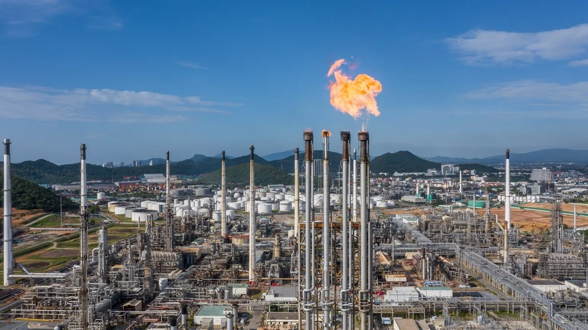 An industrial oil refinery with multiple tall smokestacks emitting flames and smoke, set against a backdrop of mountains and cityscape.
