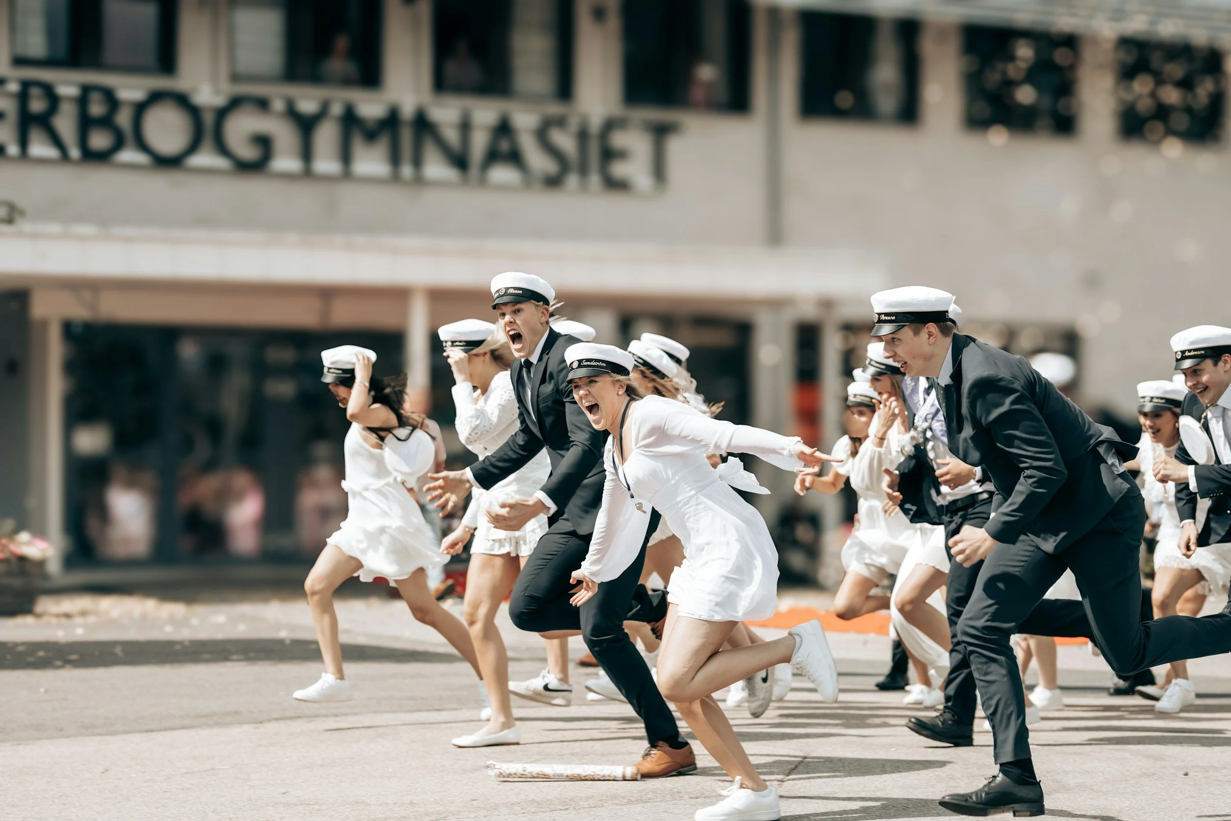 Students in white and black graduation caps, some in white dresses and some in black suits, running and celebrating outdoors on a street during graduation day.