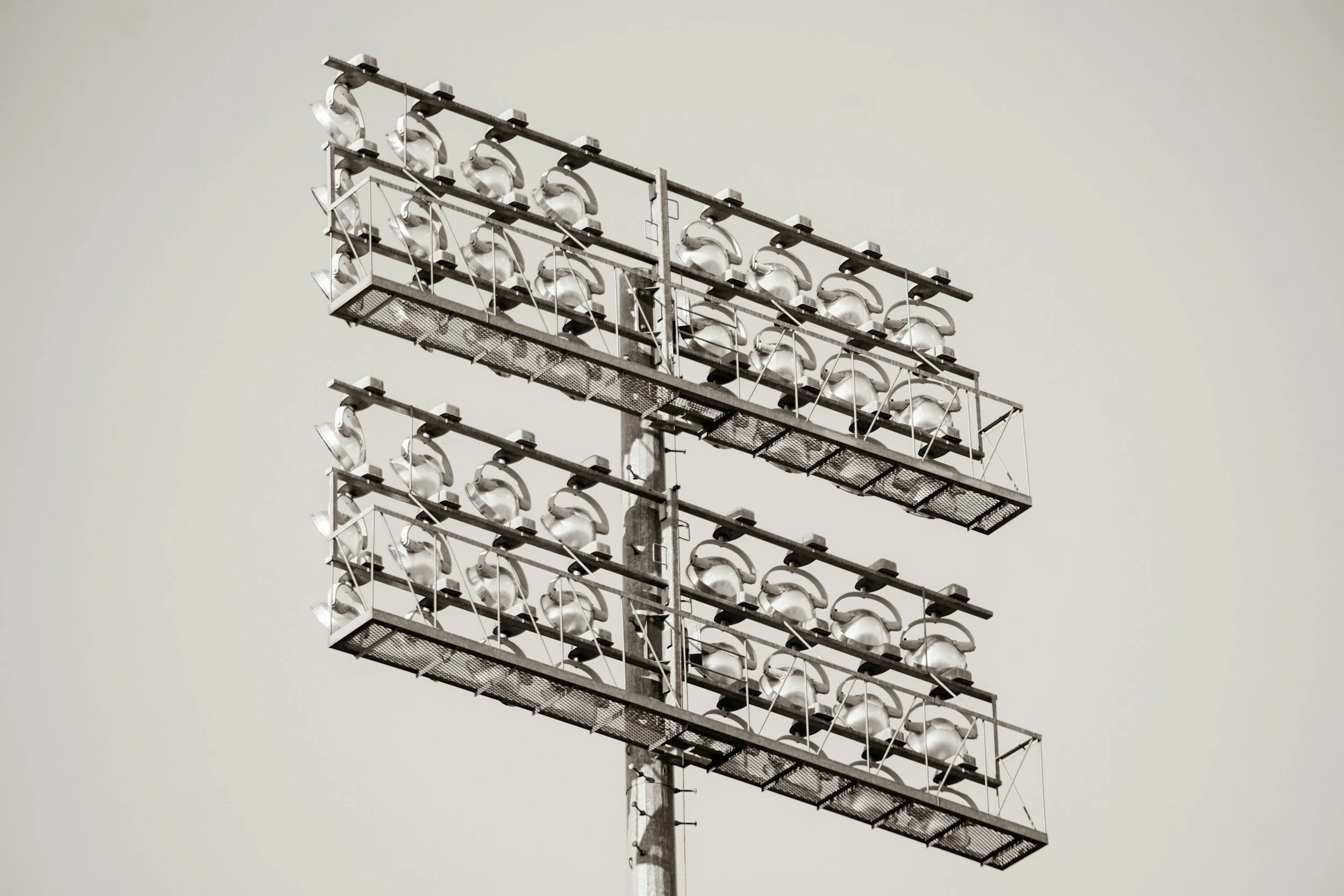 A tall stadium floodlight tower with large metal lights arranged in three horizontal rows against a clear sky.