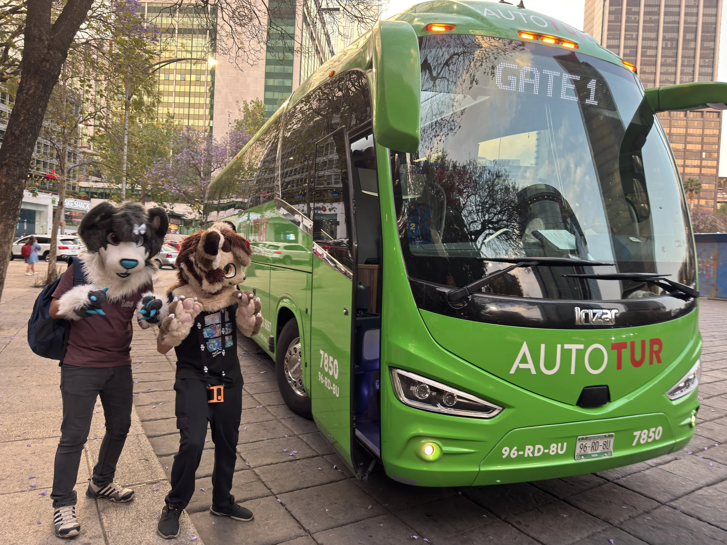 Dos personajes disfrazados de mascotas caminando cerca de un autobús verde en la ciudad, con edificios altos y árboles de fondo.