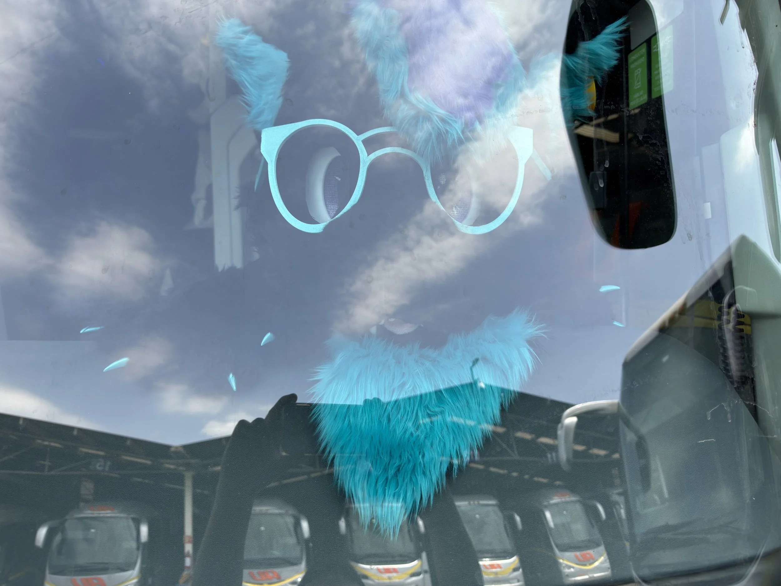 Reflejo en el parabrisas de un auto mostrando un gato de peluche azul y unas gafas de sol. Al fondo, nubes en el cielo y otros vehículos en un estacionamiento.