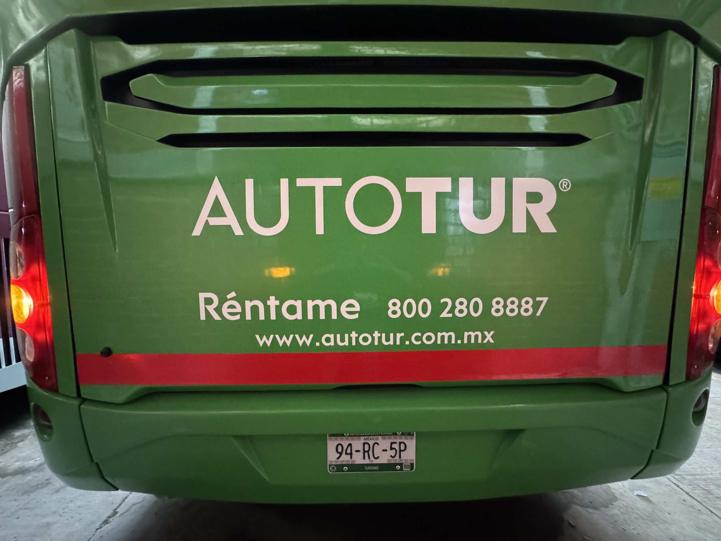 Furgoneta verde con anuncios de autobús que promocionan una empresa de tours con teléfono y sitio web en español.