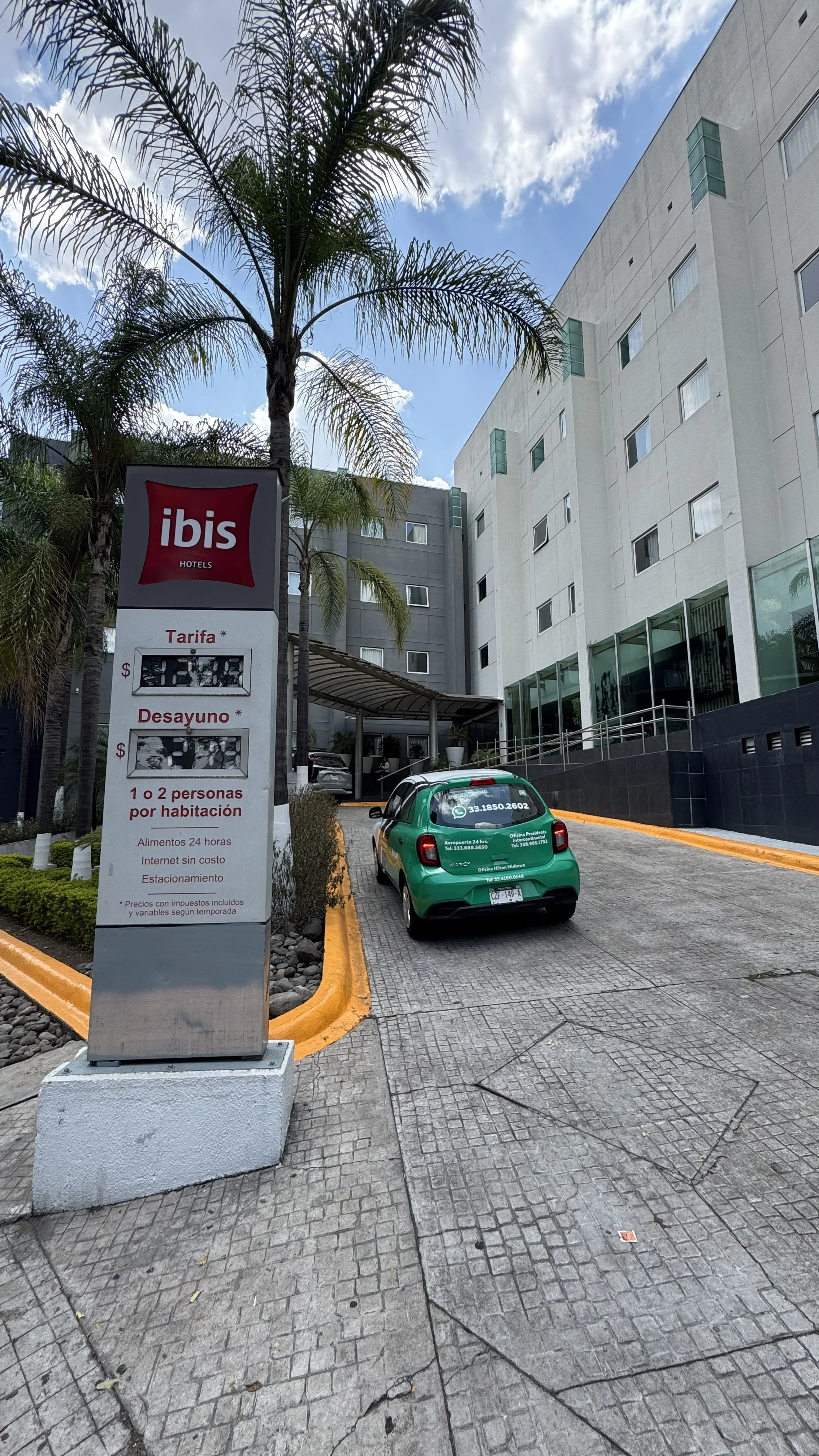 Entrada de un hotel ibis con señalización de tarifas y servicios, vehículos estacionados en la entrada del hotel, edificios modernos y palmeras en el fondo, cielo parcialmente nublado.