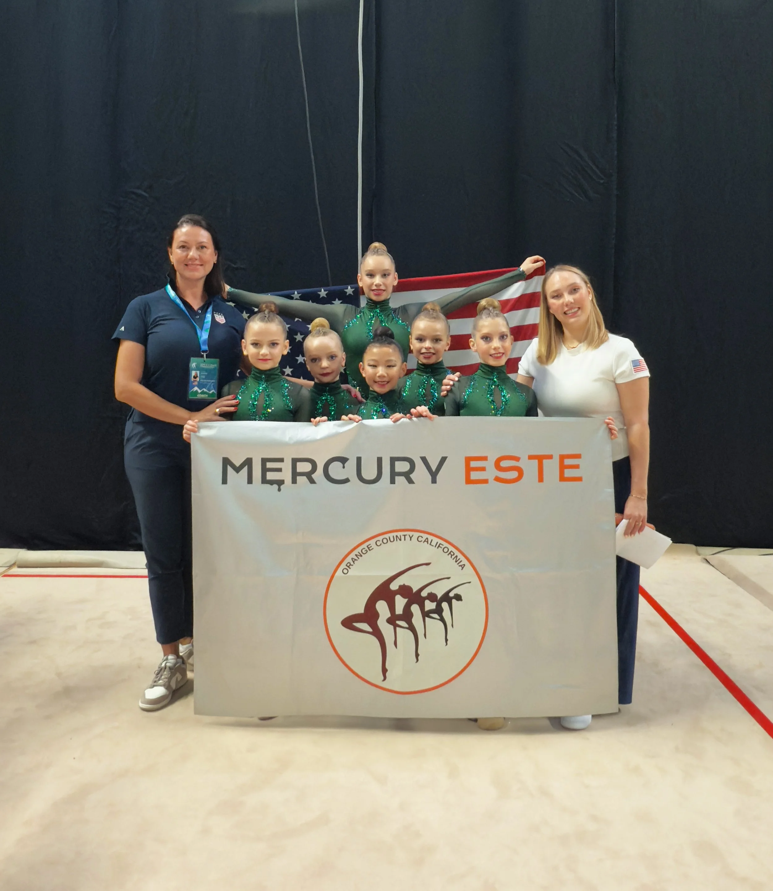 Group of young artistic gymnasts holding a banner that reads 'Mercury Este' with the logo of Orange County, California, in front of a large American flag backdrop.