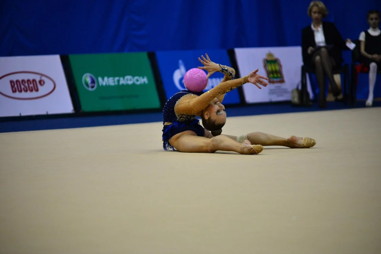 A young gymnast performing a floor routine with a pink ball, sitting on the mat with legs spread and arms extended.