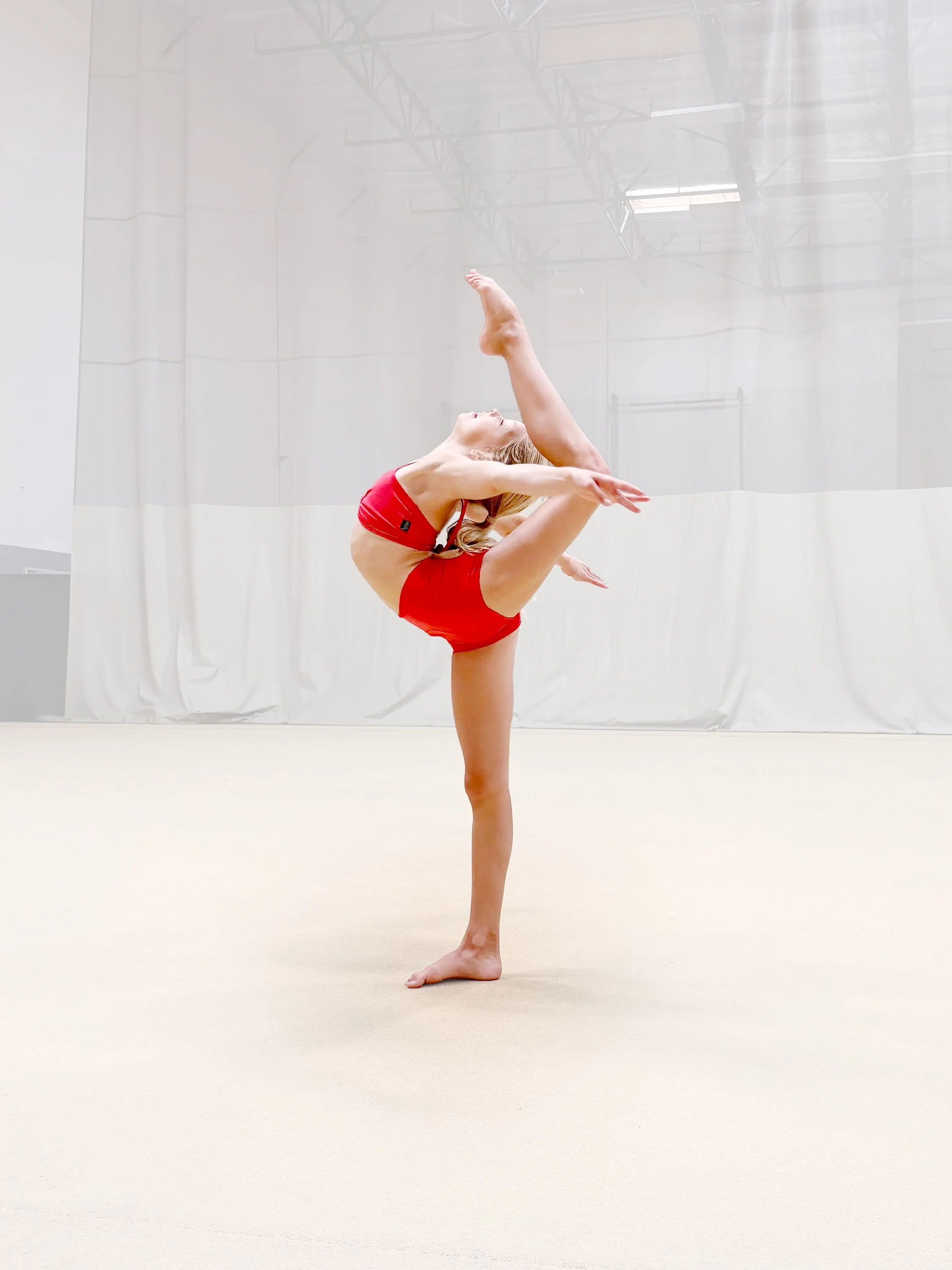 A female gymnast in a red outfit is performing a balance pose on a practice mat in an indoor gymnasium.