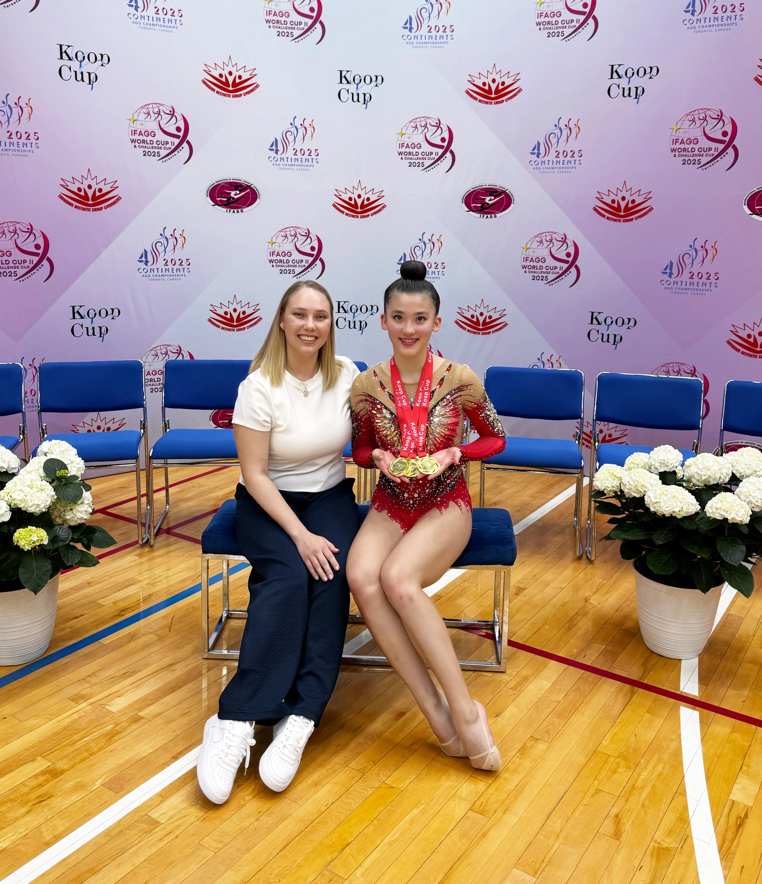 A young female gymnast in a red and gold leotard sitting on a bench, holding three gold medals, with a woman beside her, both smiling, in an indoor sports venue with blue chairs, white flowers, and event banners in the background.